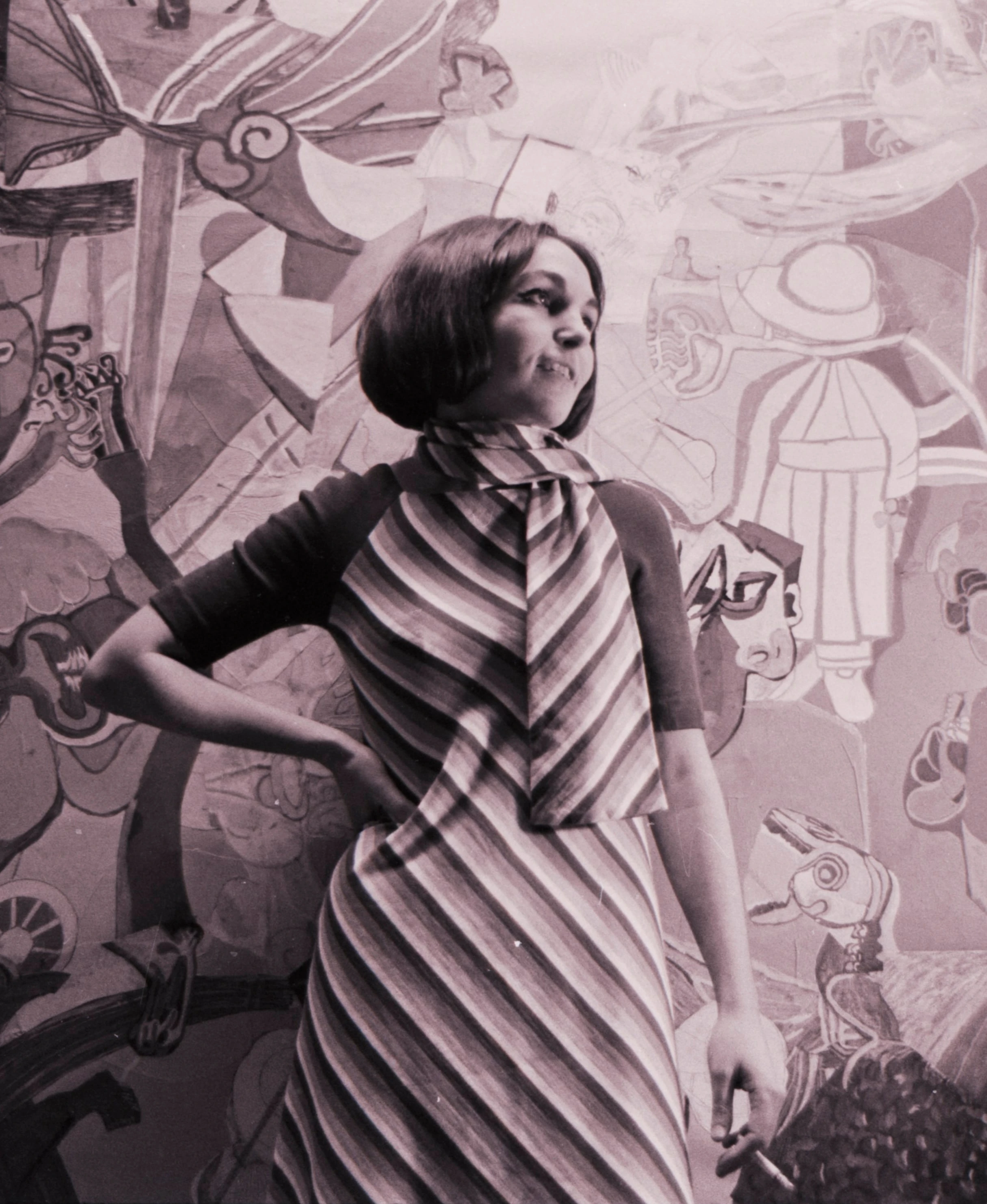 Black and white photograph of a young woman (Paula Rego). She is wearing a striped dress and has her right hand on her hip while looking to the side.