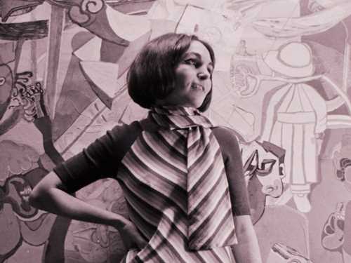 Black and white photograph of a young woman (Paula Rego). She is wearing a striped dress and has her right hand on her hip while looking to the side.