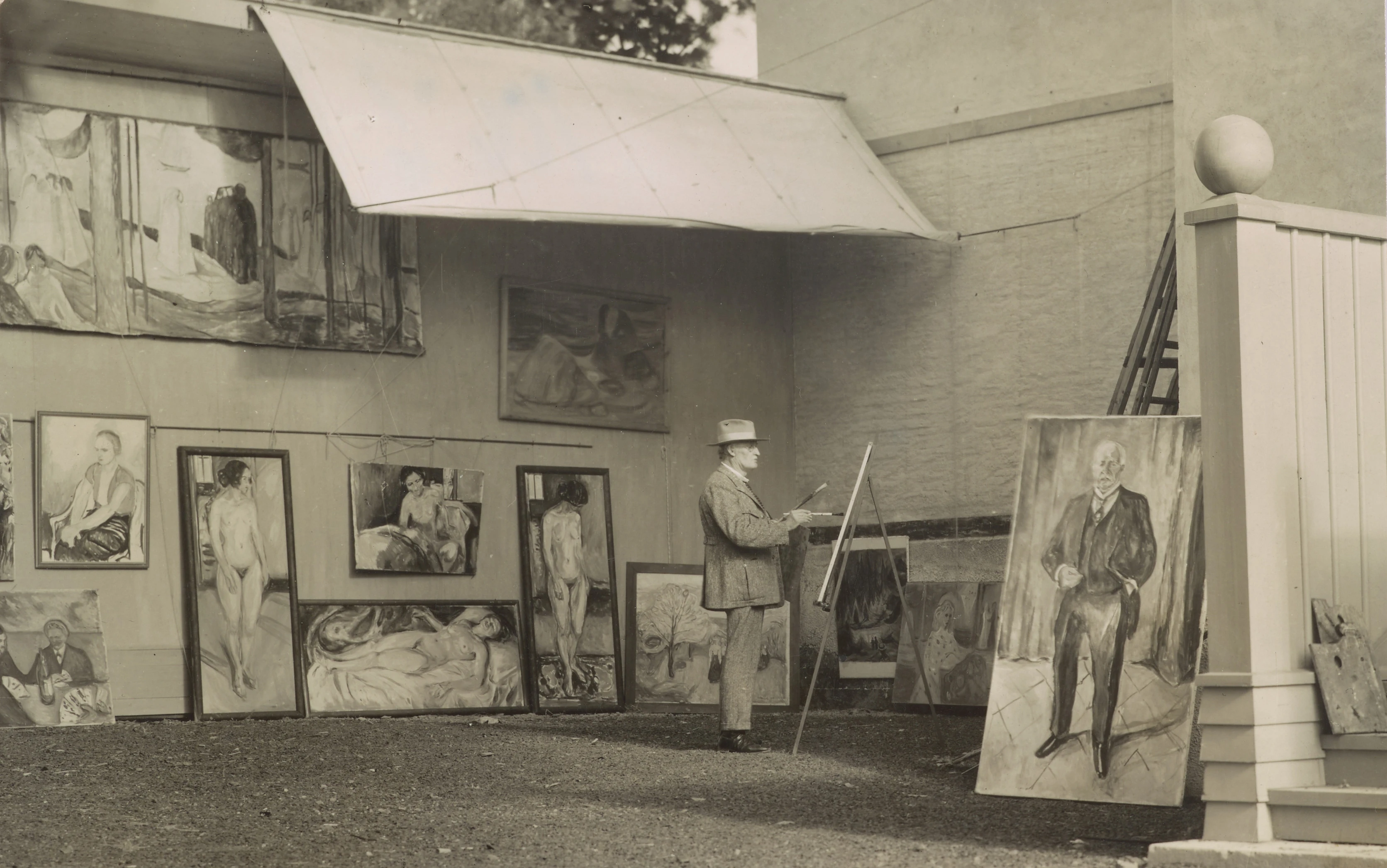 Edvard Munch is standing and painting in his open-air studio at Ekely. He is surrounded by his own works and is wearing a hat to shield himself from the sun and a light-coloured suit.
