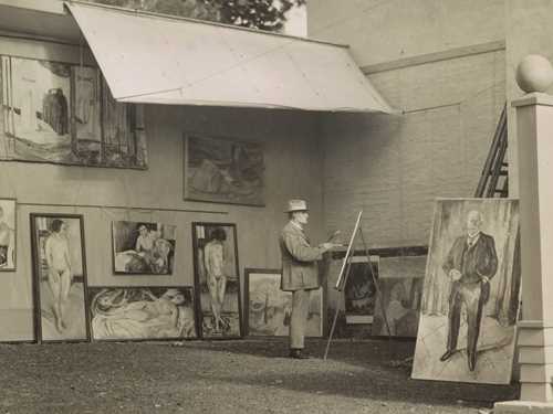 Edvard Munch is standing and painting in his open-air studio at Ekely. He is surrounded by his own works and is wearing a hat to shield himself from the sun and a light-coloured suit.
