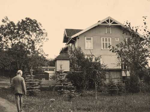 Edvard Munch walks along the road leading up to Ekely. There is a grassy meadow and trees in front of the house.