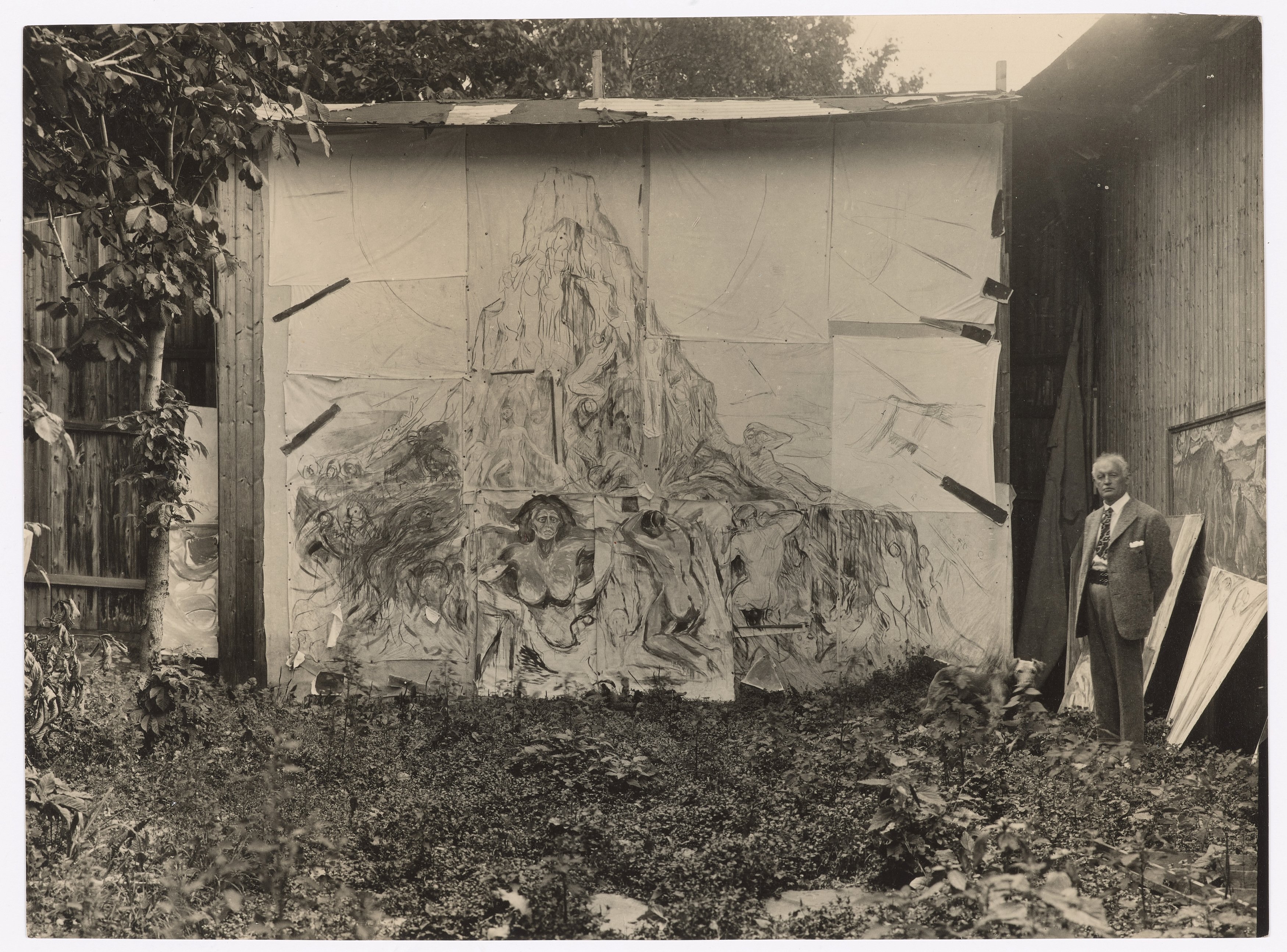 Munch’s open-air studio at Ekely. Edvard Munch is seen on the right side of the image, standing in front of the painting The Human Mountain. Photograph.