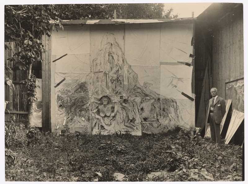 Munch’s open-air studio at Ekely. Edvard Munch is seen on the right side of the image, standing in front of the painting The Human Mountain. Photograph.