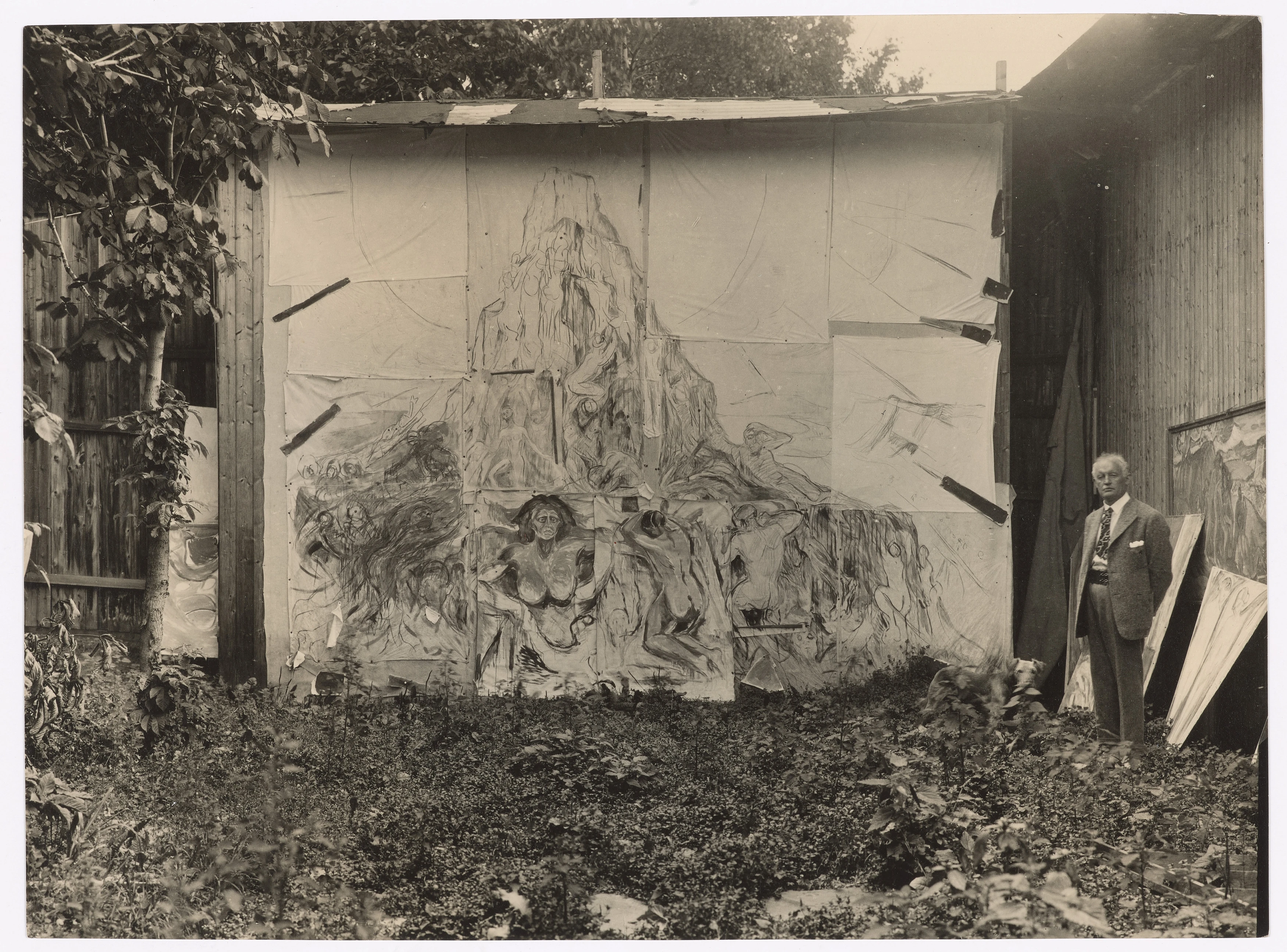 Munchs friluftsatelier på Ekely. Vi ser Edvard Munch til høyre i bildet. Han står foran maleriet Menneskeberget. Fotografi