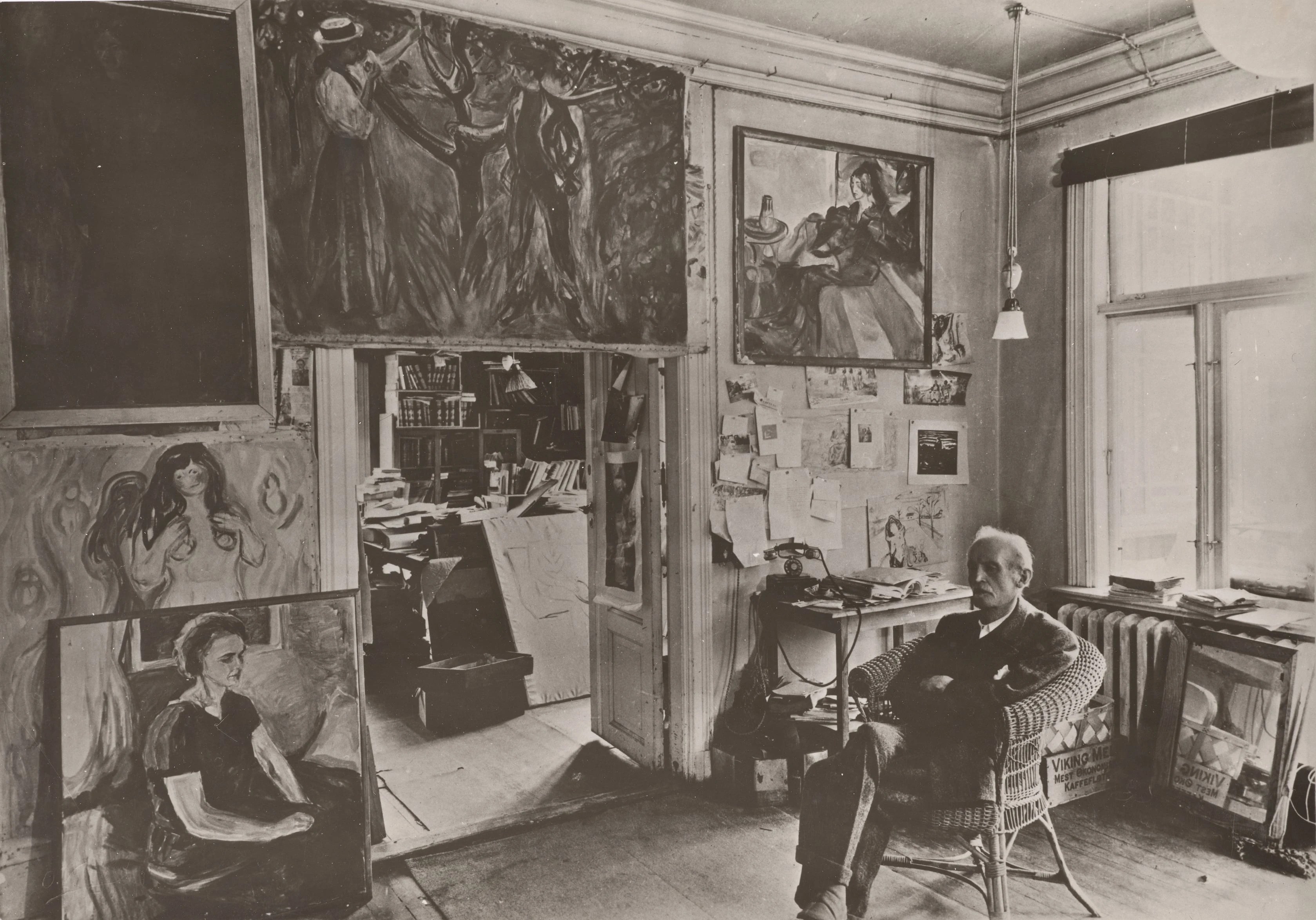 Edvard Munch sits in a wicker chair in a room at Ekely, surrounded by his paintings. Photograph.
