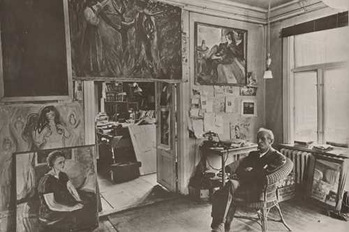 Edvard Munch sits in a wicker chair in a room at Ekely, surrounded by his paintings. Photograph.