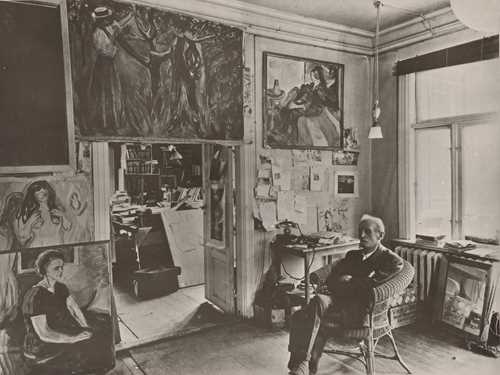 Edvard Munch sits in a wicker chair in a room at Ekely, surrounded by his paintings. Photograph.