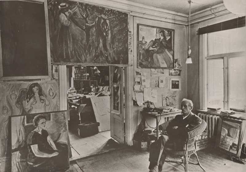 Edvard Munch sits in a wicker chair in a room at Ekely, surrounded by his paintings. Photograph.