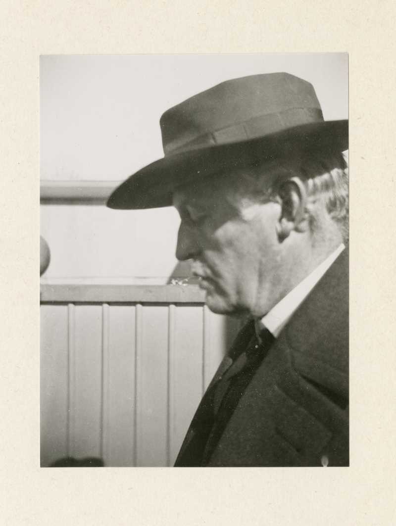 Et svart-hvitt fotografi av Edvard Munchs ansikt i profil. Han har på seg dressjakke, og en mørk hatt som kaster skygge over øynene, som er lukket.