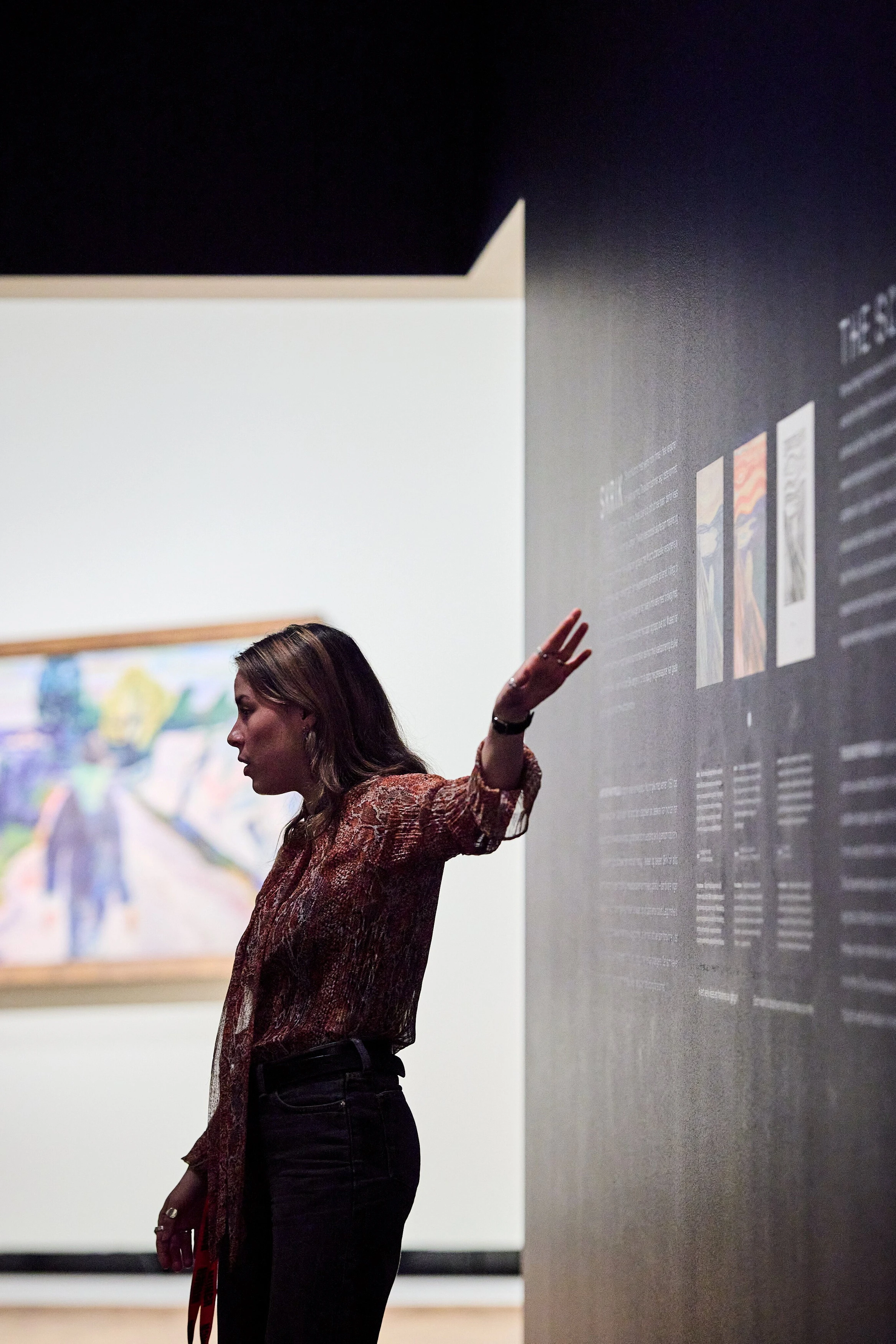 A mediator at MUNCH stands in front of a blue wall with text explaining the different versions of The Scream held by the Munch Museum. Photograph.
