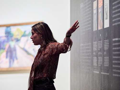 A mediator at MUNCH stands in front of a blue wall with text explaining the different versions of The Scream held by the Munch Museum. Photograph.