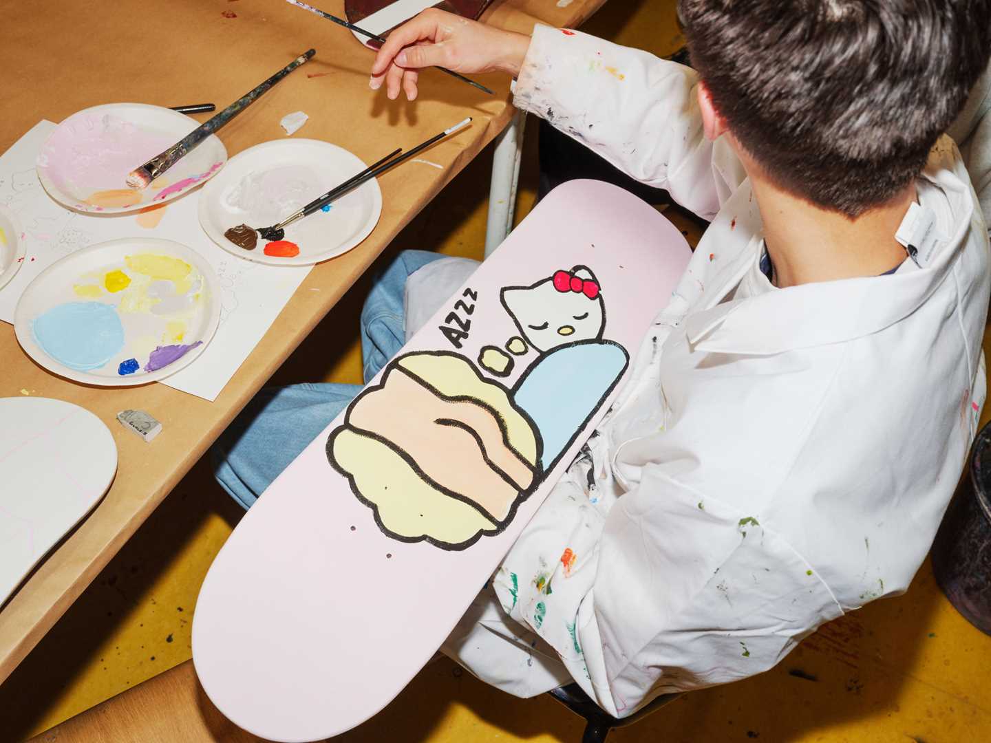 A boy sits painting on the underside of a skateboard. Photograph.