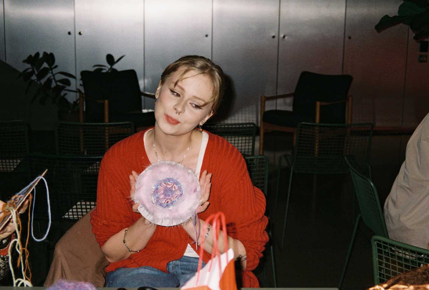 A girl wearing a red sweater sits, showing a handicraft she has made. Photograph.