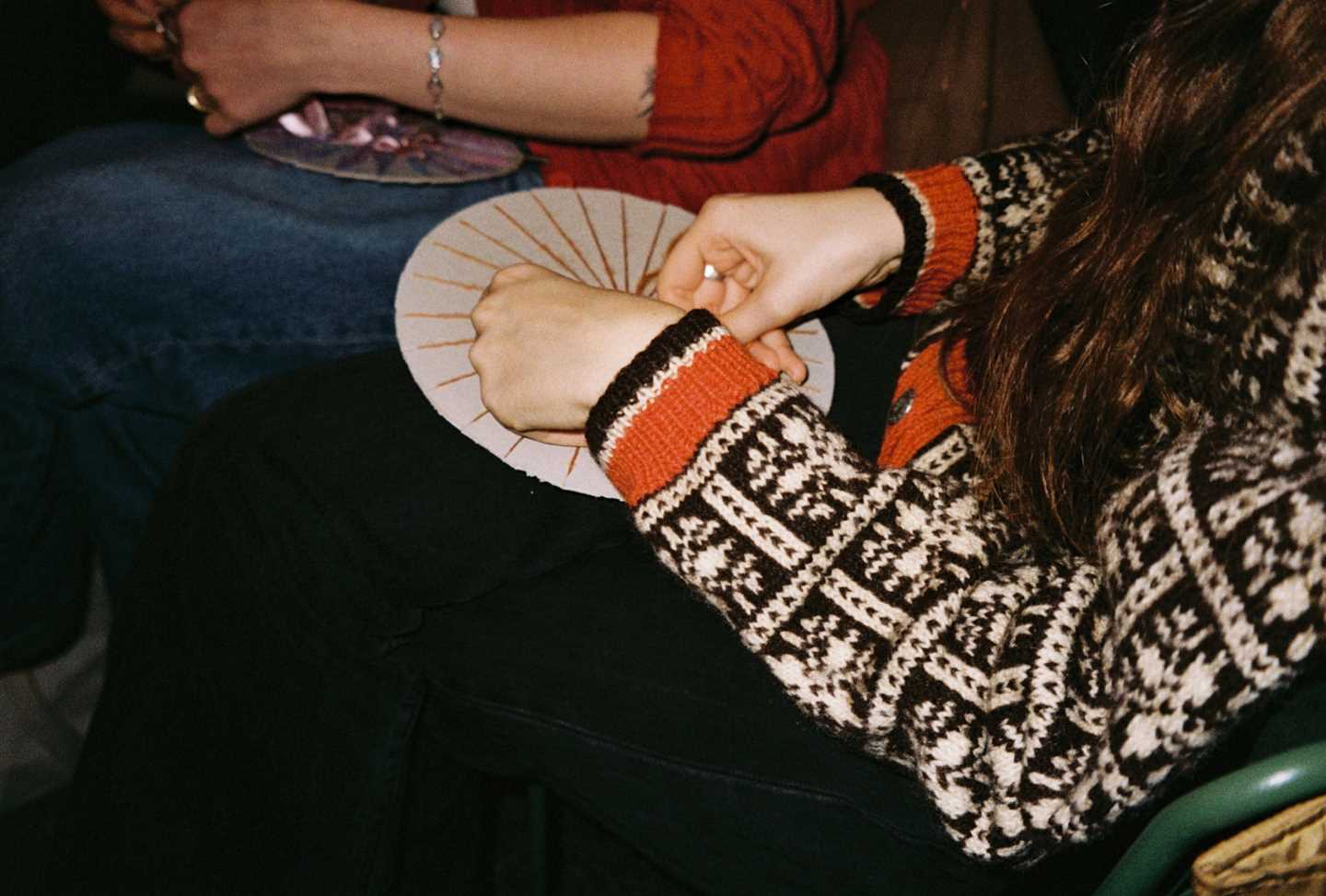 A girl in a wool sweater sits working on a handicraft. Photograph.
