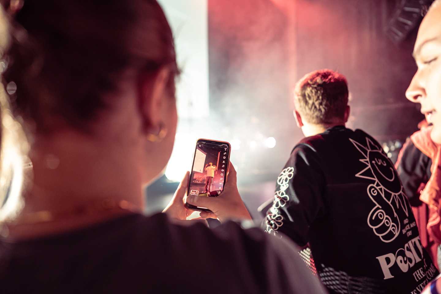Young people at a concert. One is taking a photo of the artist on stage with a mobile phone. Photograph.