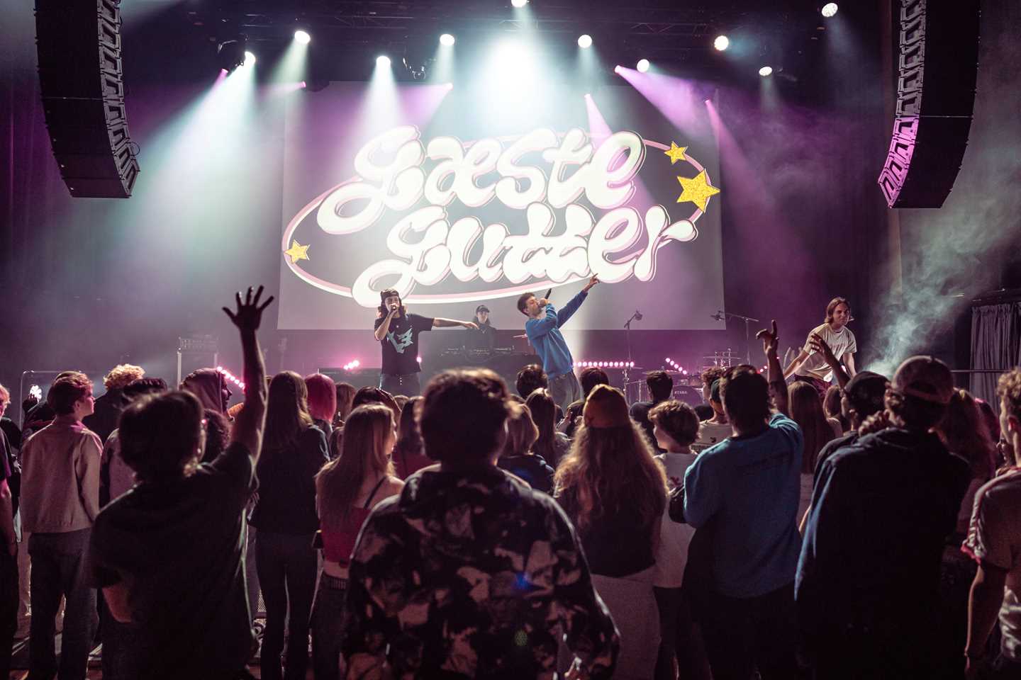 A packed concert hall with people standing with their backs to the camera, watching a stage where several hip-hop artists are performing. Purple stage lights illuminate the scene, and a backdrop behind the stage reads “Gæste gutter.” Photograph.