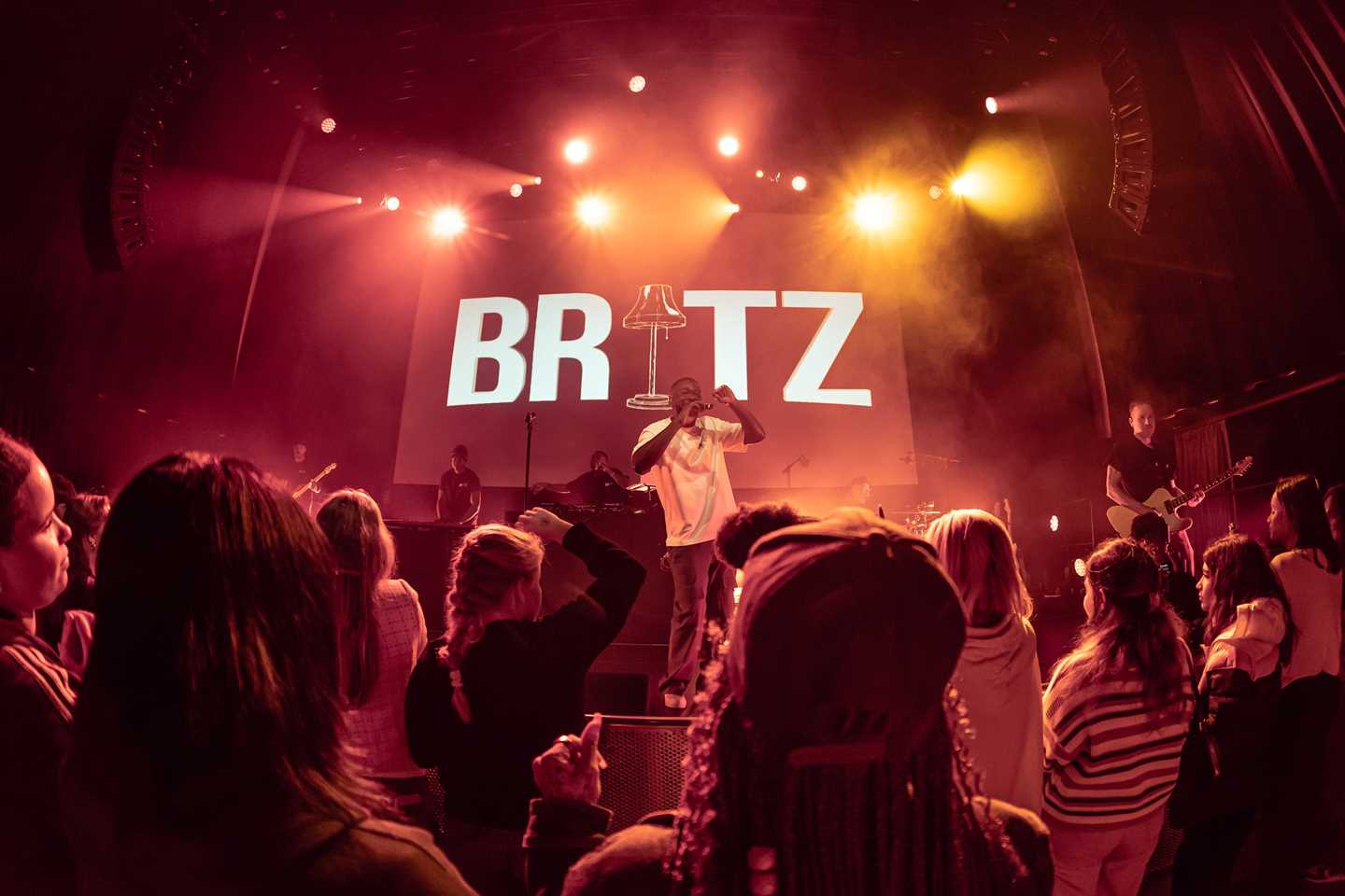 A dark concert hall with red stage lighting, a large audience, and an artist performing on stage. Photograph.