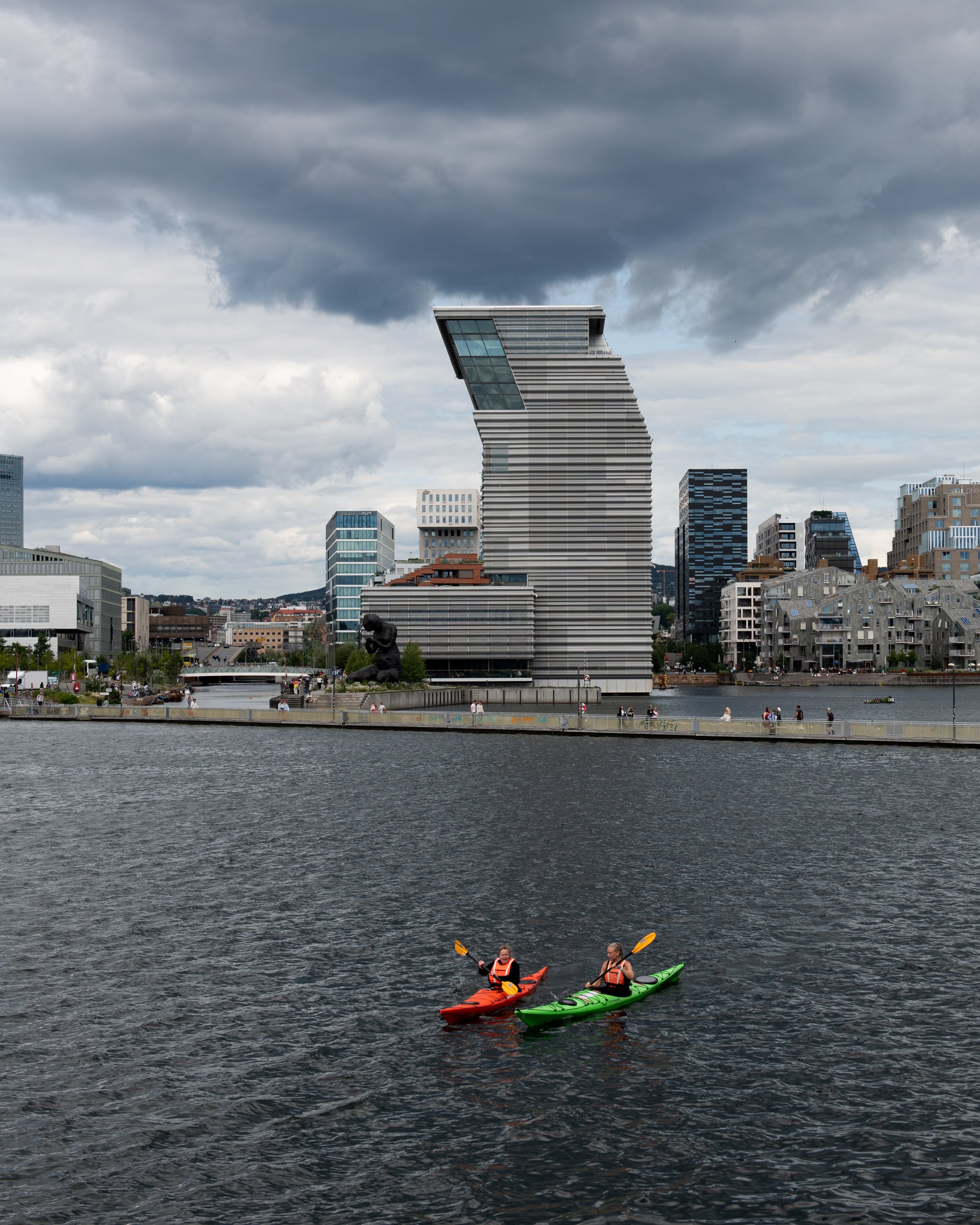 MUNCH seen from Sørenga. In the foreground, two people paddle kayaks, with a floating walkway behind them. The sky is blue-grey and cloudy. Photograph.