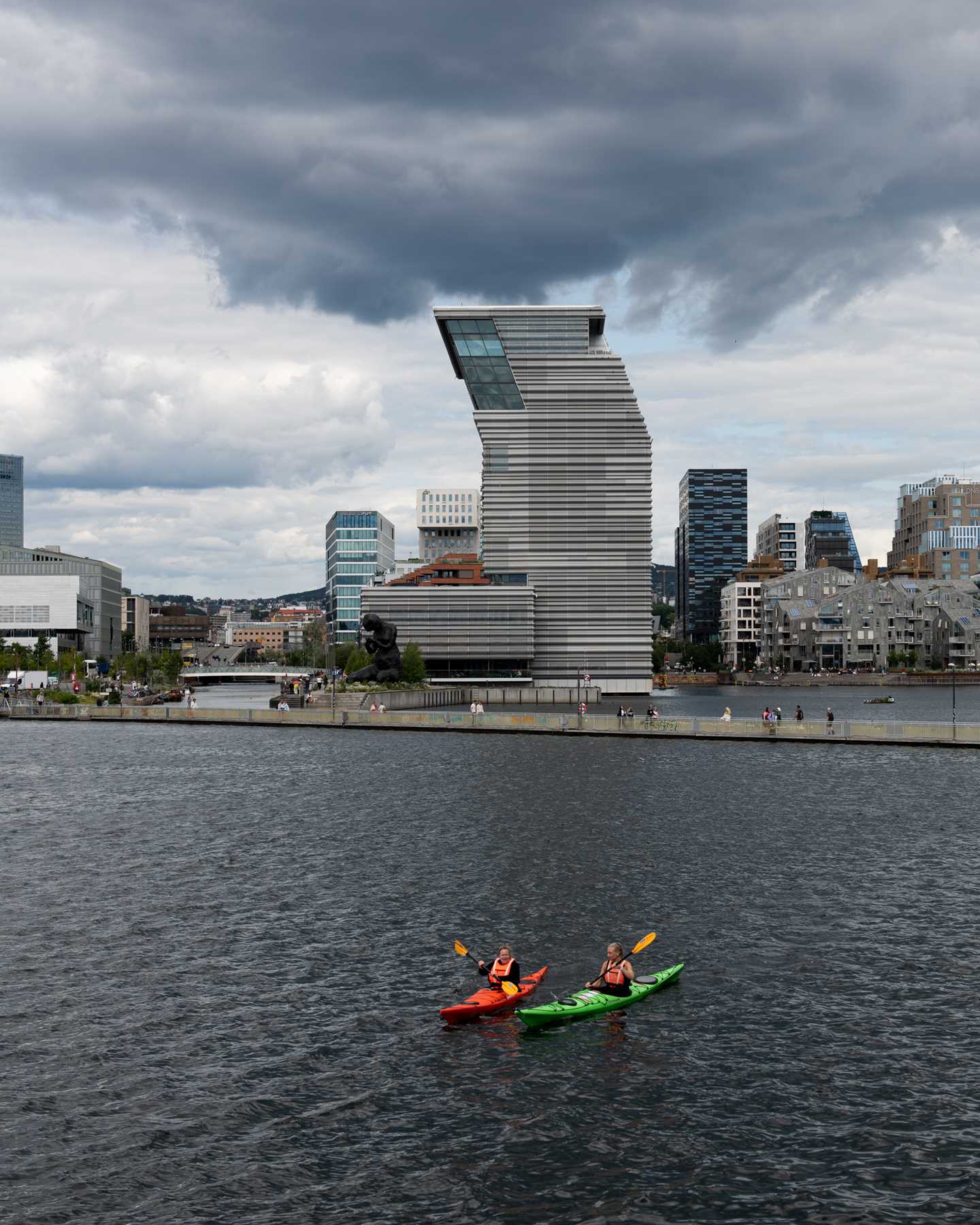 MUNCH seen from Sørenga. In the foreground, two people paddle kayaks, with a floating walkway behind them. The sky is blue-grey and cloudy. Photograph.