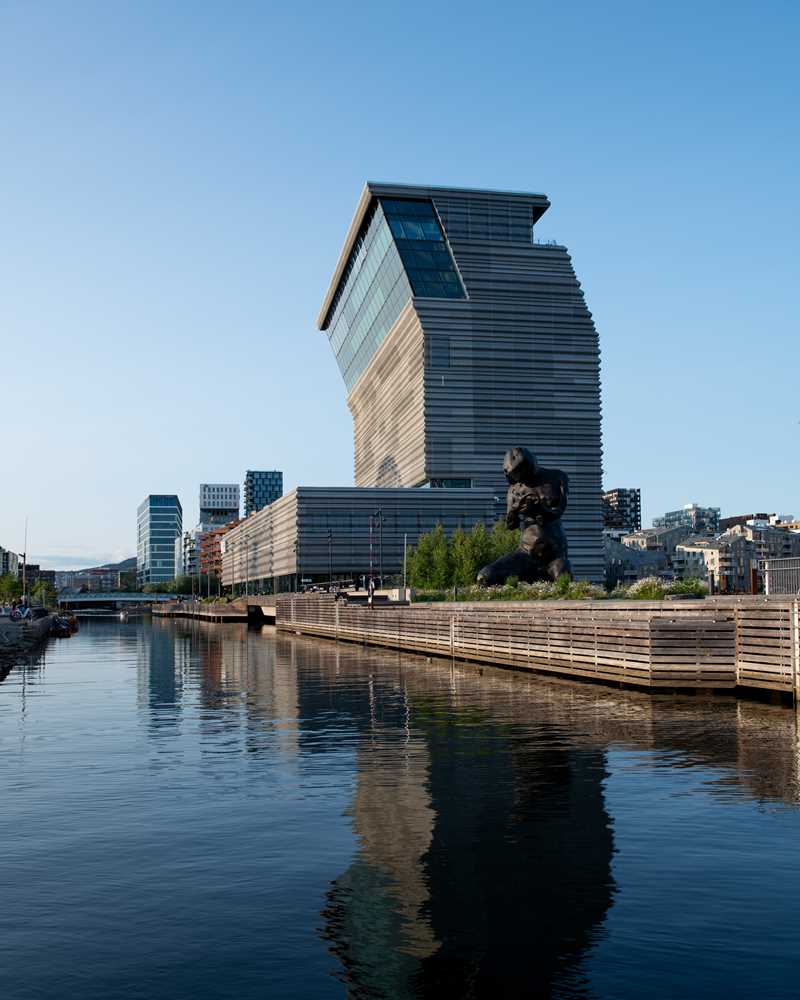 The MUNCH building seen from the south side in bright sunlight, reflected in the water, with the sculpture The Mother in the foreground. Photograph.