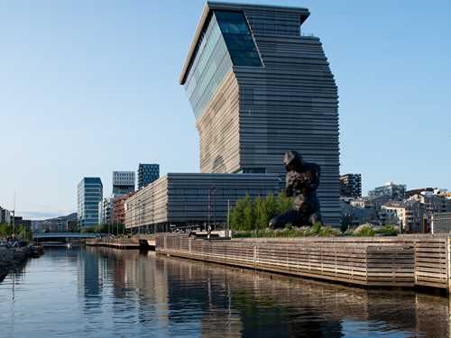 The MUNCH building seen from the south side in bright sunlight, reflected in the water, with the sculpture The Mother in the foreground. Photograph.