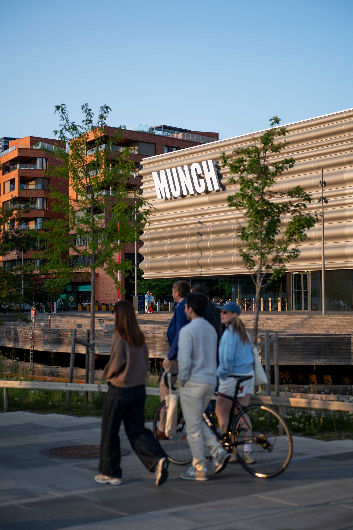 Part of the MUNCH building seen from the west. In the foreground, a group of four friends walk with a bicycle, and in the background, the building with the MUNCH logo is framed between two green trees. Photograph.