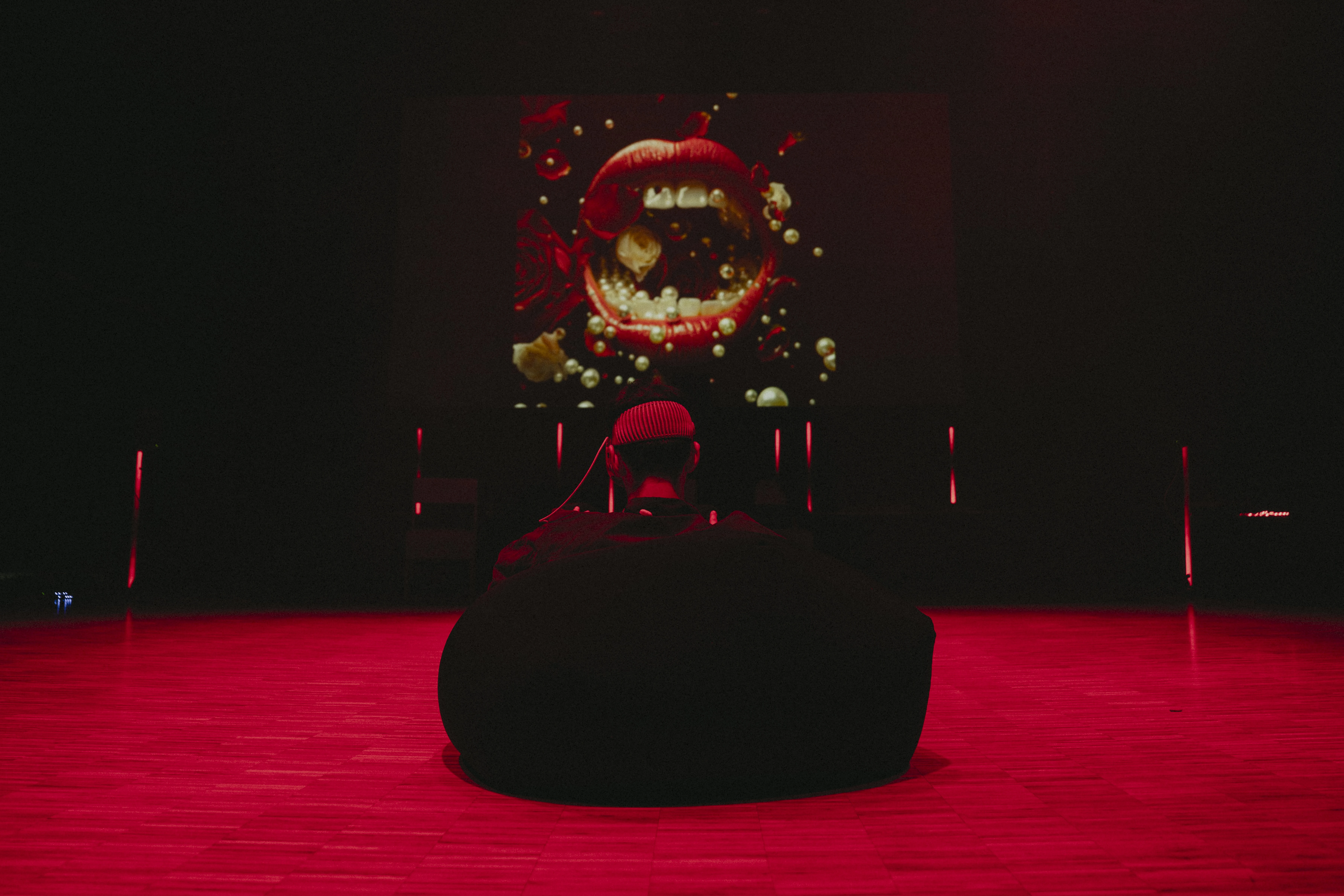 A person sits with their back to the camera in a beanbag chair on the floor. The room is lit so that the floor appears red. On the wall in front of the person is an image of a large, open mouth with red lips and white teeth. Photograph.