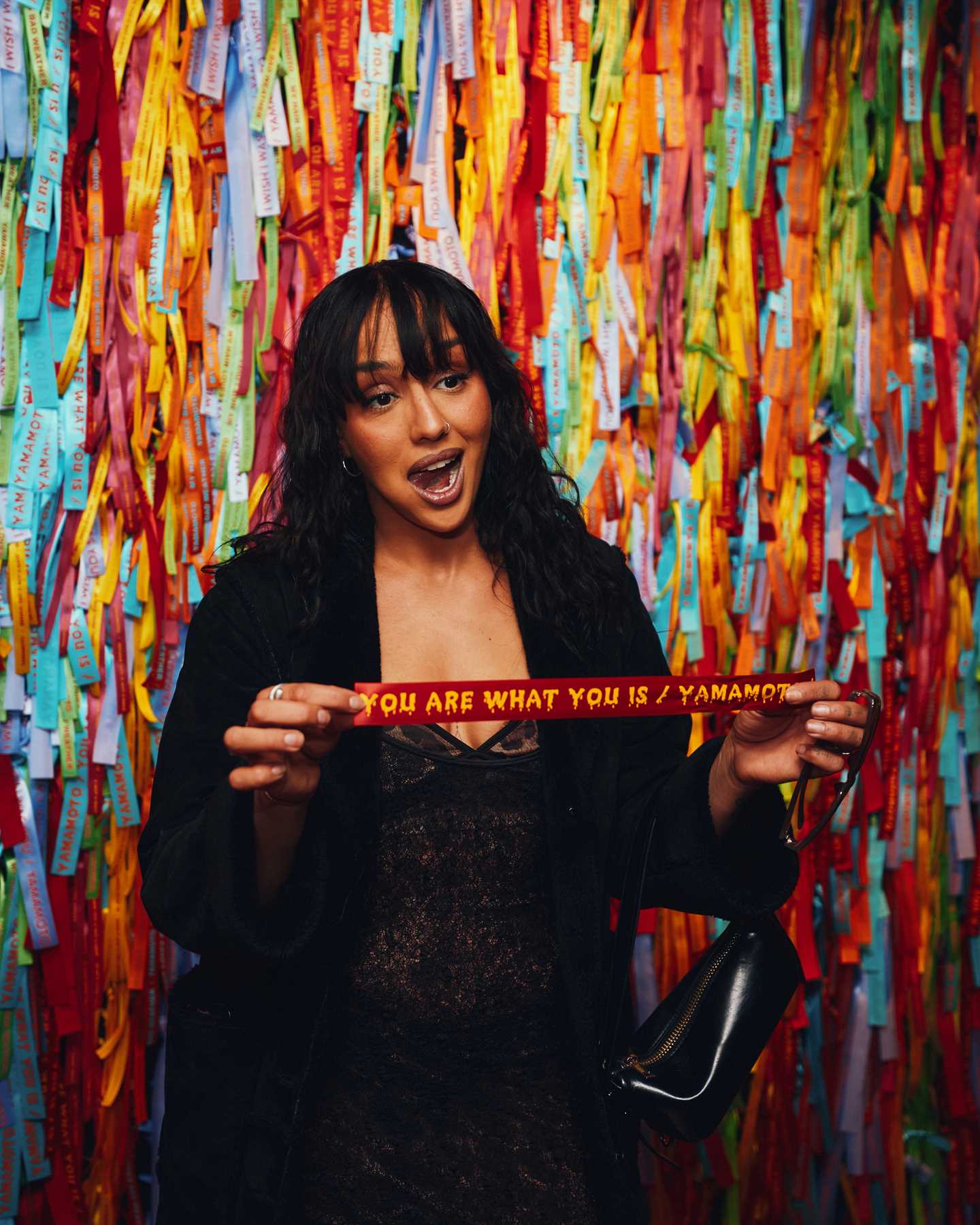 A woman stands in front of a wall of silk ribbons, holding one she has taken down. Photograph.