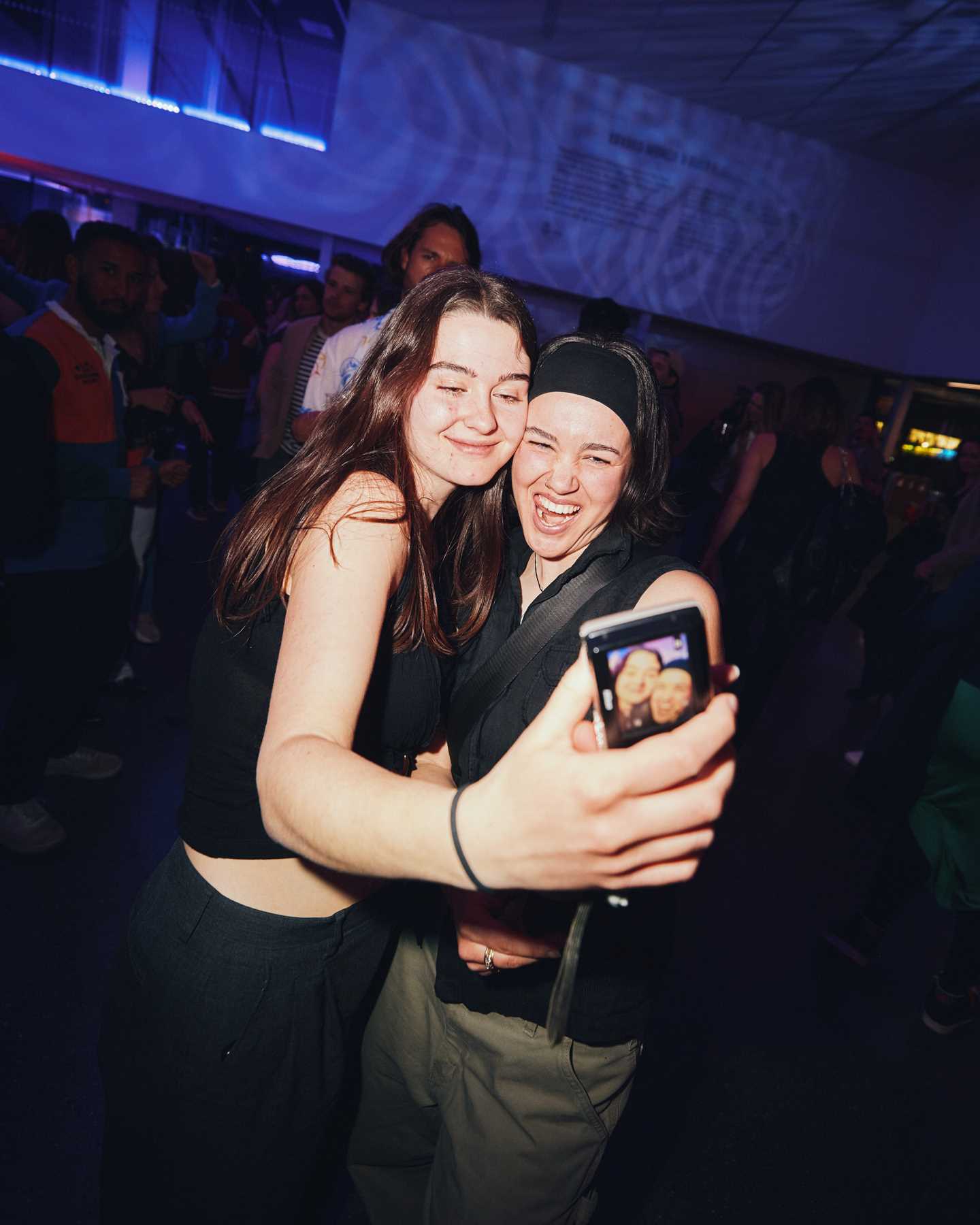 Two friends on the dance floor embracing, one taking a photo with her phone. Both are looking at the phone camera. Photograph.