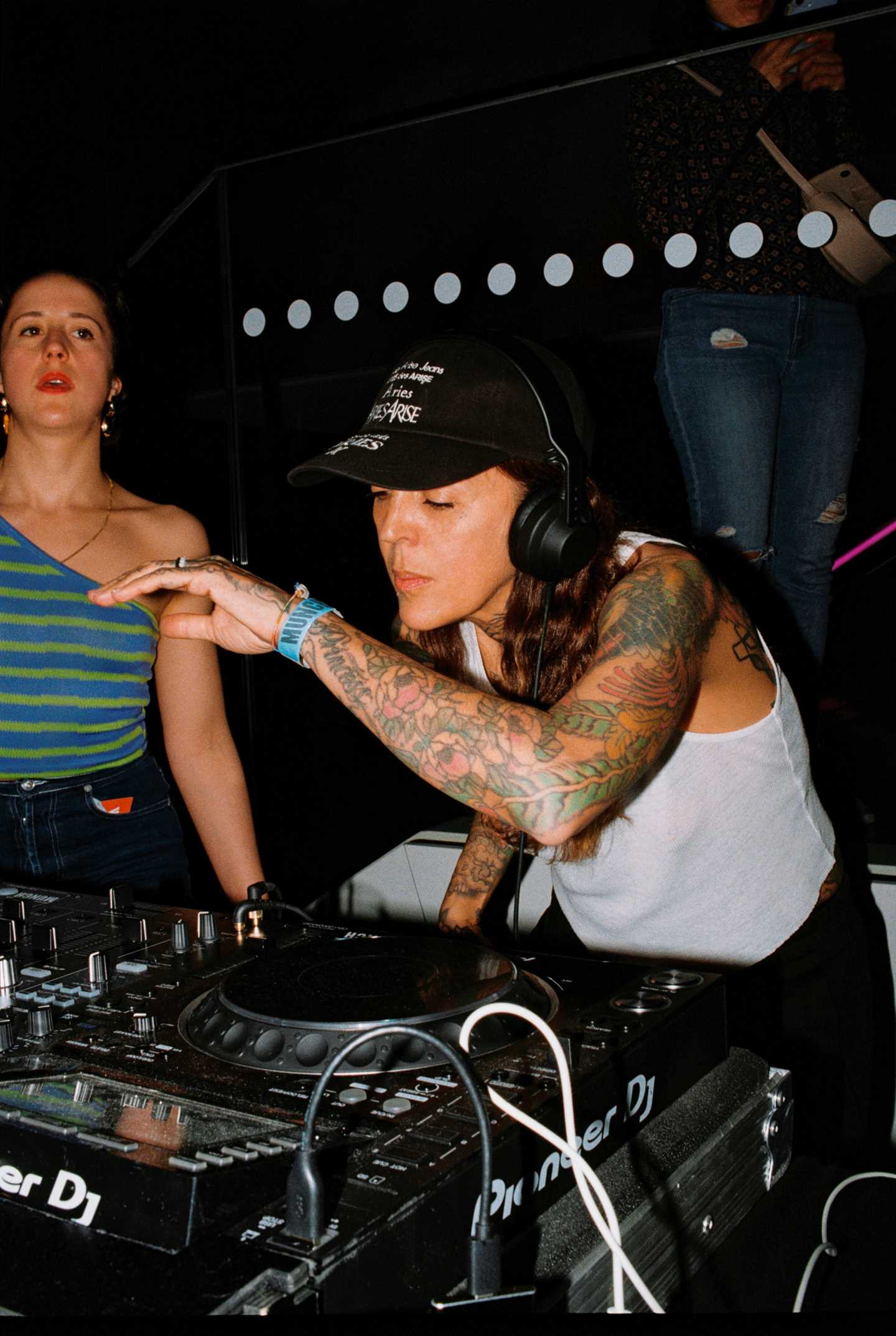 A DJ wearing headphones stands behind a mixing console, spinning records. Photograph.