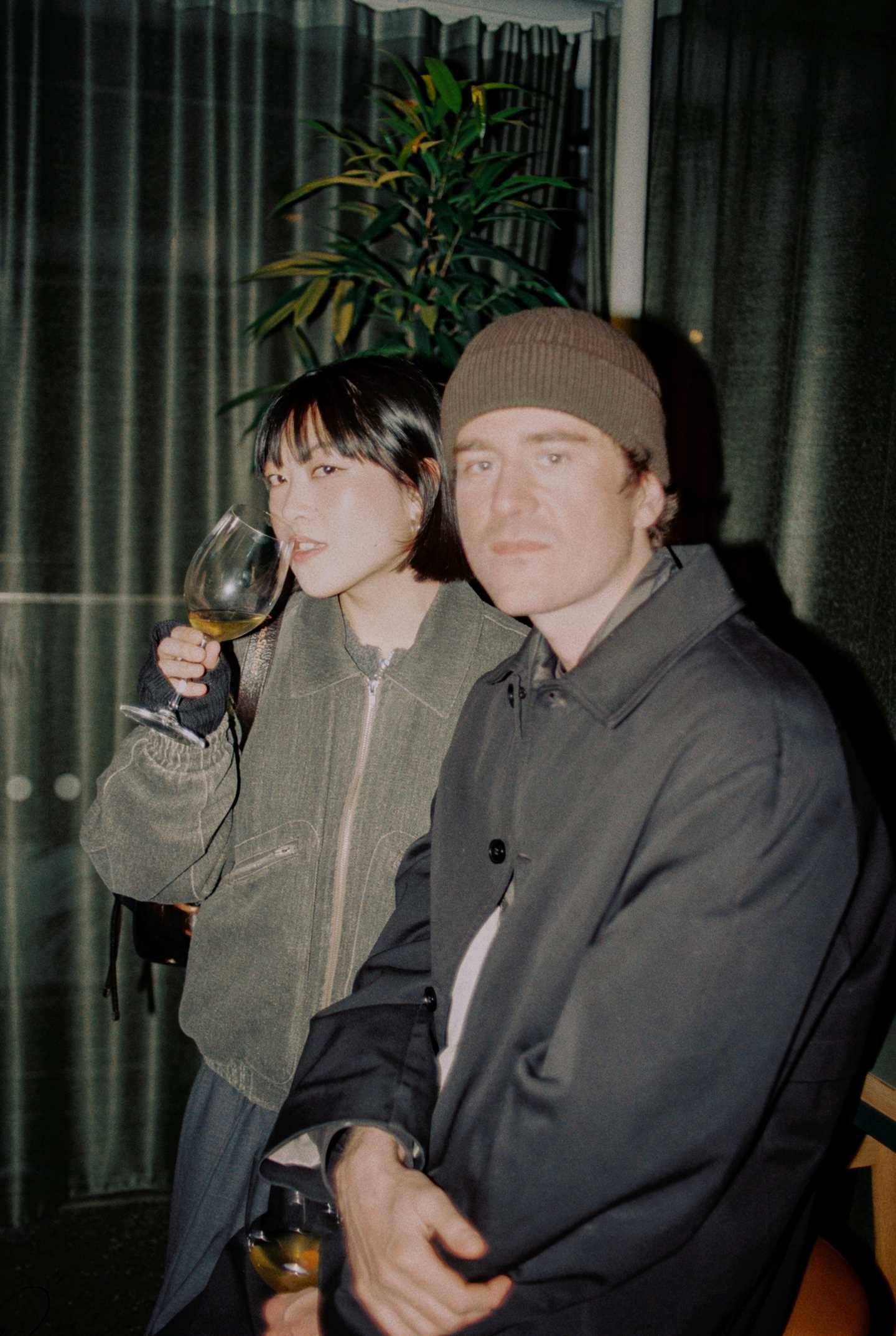 A girl and a boy sit at the bar in MUNCH, each with a glass of white wine, looking directly at the camera. Photograph.