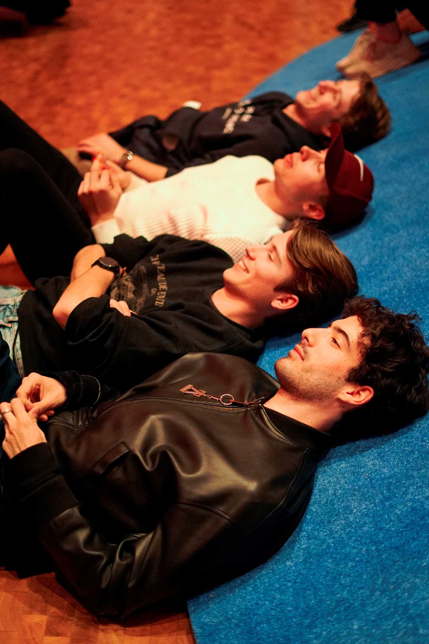Four boys lying on the floor, leaning against a gentle slope covered in blue fabric. Photograph.