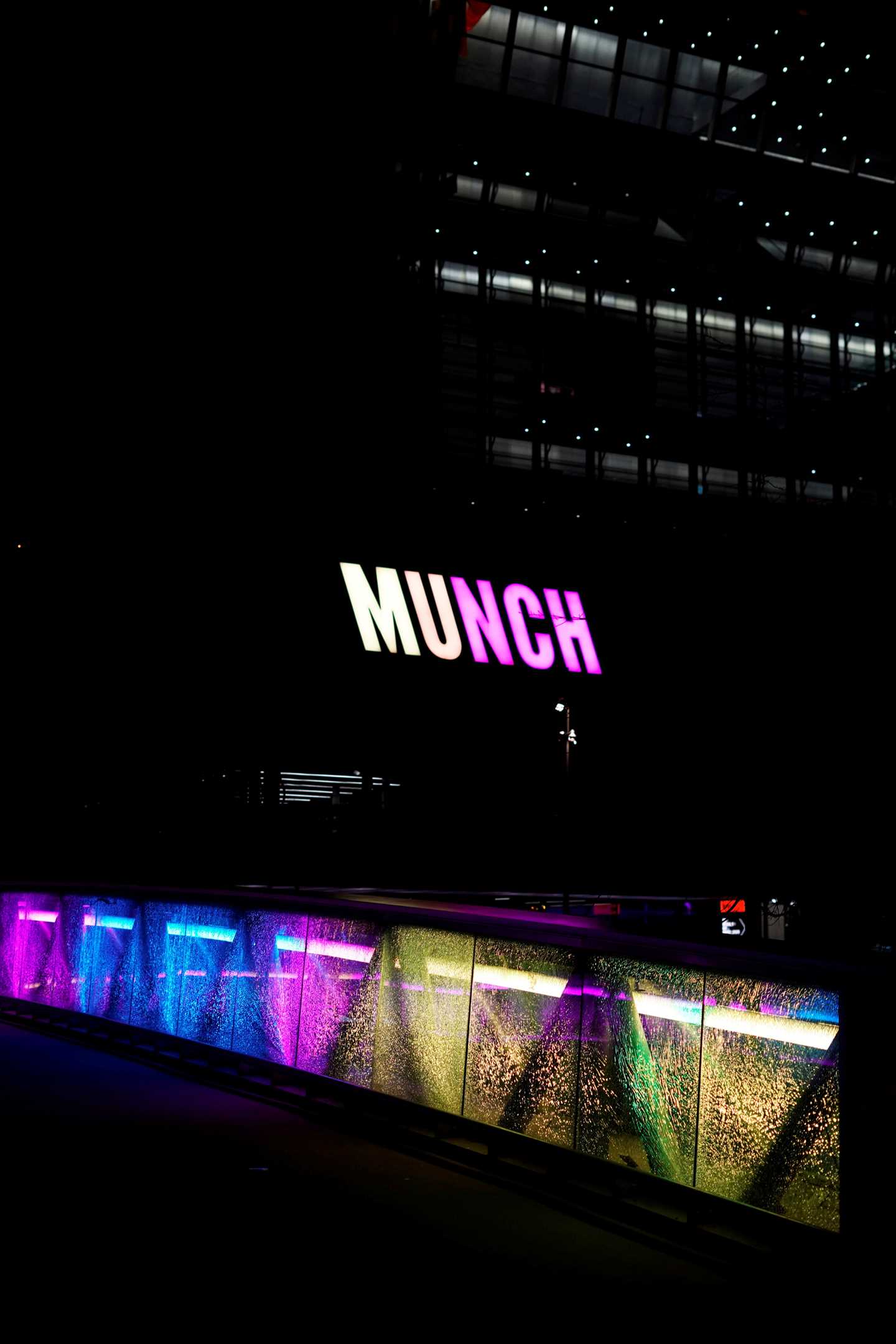 The façade of the MUNCH building at night. The logo glows in yellow and pink, as does the bridge in front of the building. Photograph.