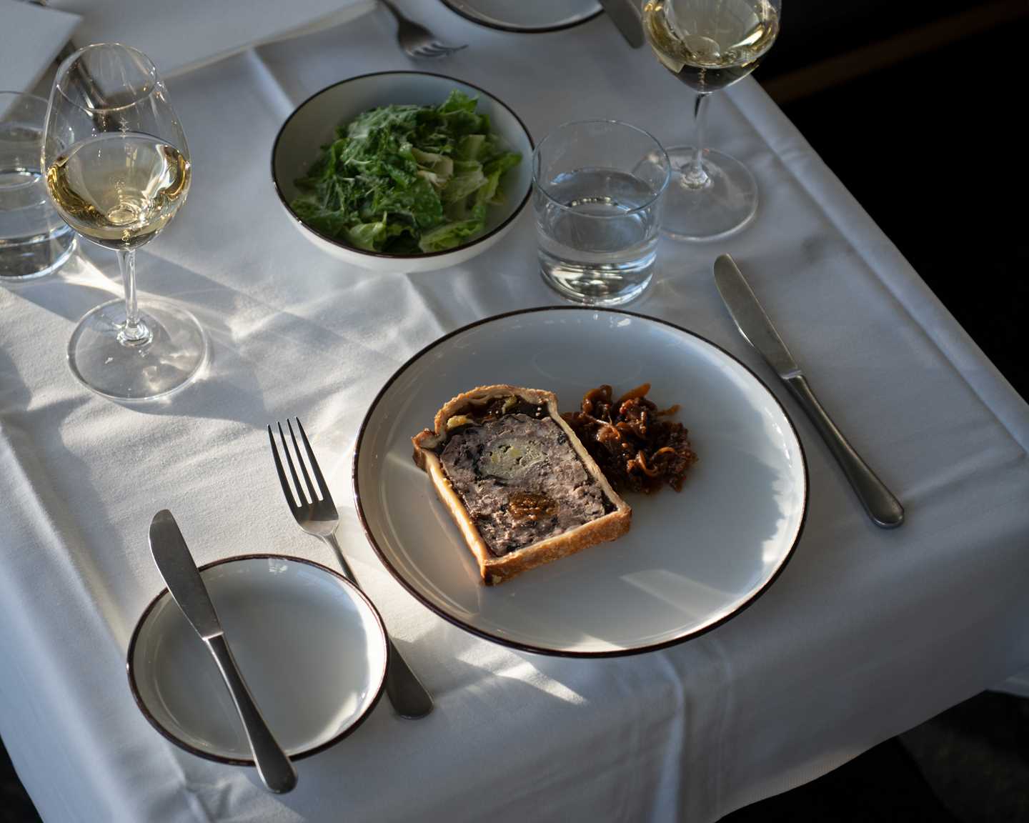 A table set for two with a white tablecloth. Two glasses of white wine, a green salad in a bowl at the centre, and a plate with pâté on one of the place settings. Photograph.