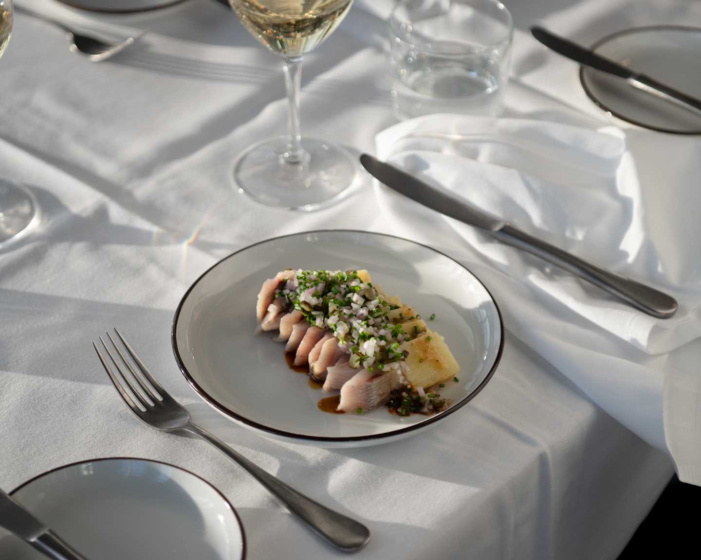 A table set with a white tablecloth and stemmed glasses. Closest to the camera is a plate of pickled herring with potatoes, elegantly arranged, beside a glass of white wine. Photograph.