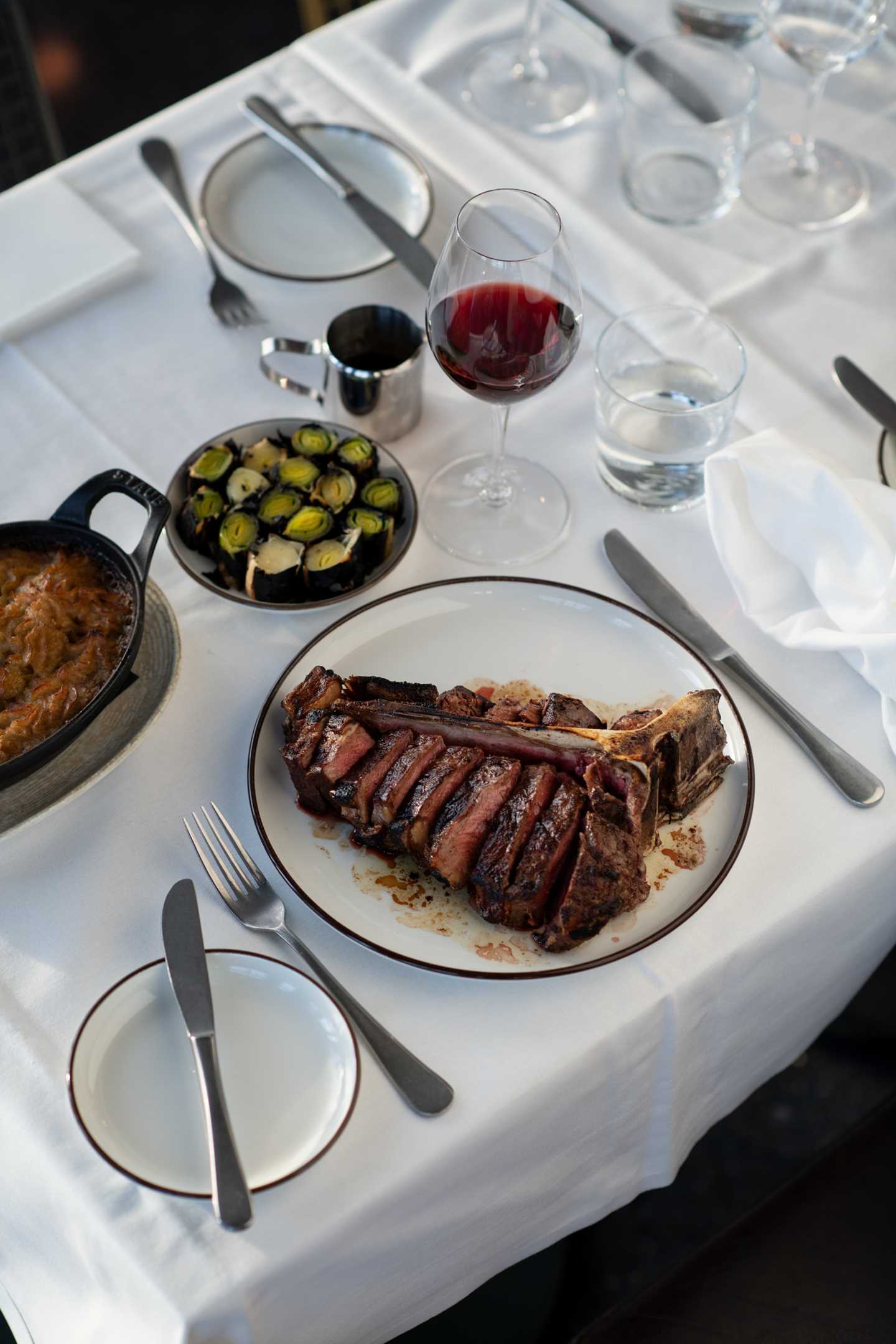 A table set with a white tablecloth, cutlery, and stemmed glasses. In focus is a plate with a hearty T-bone steak, beside a glass of red wine, a plate of roasted leeks, and a baking dish of potatoes. Photograph.
