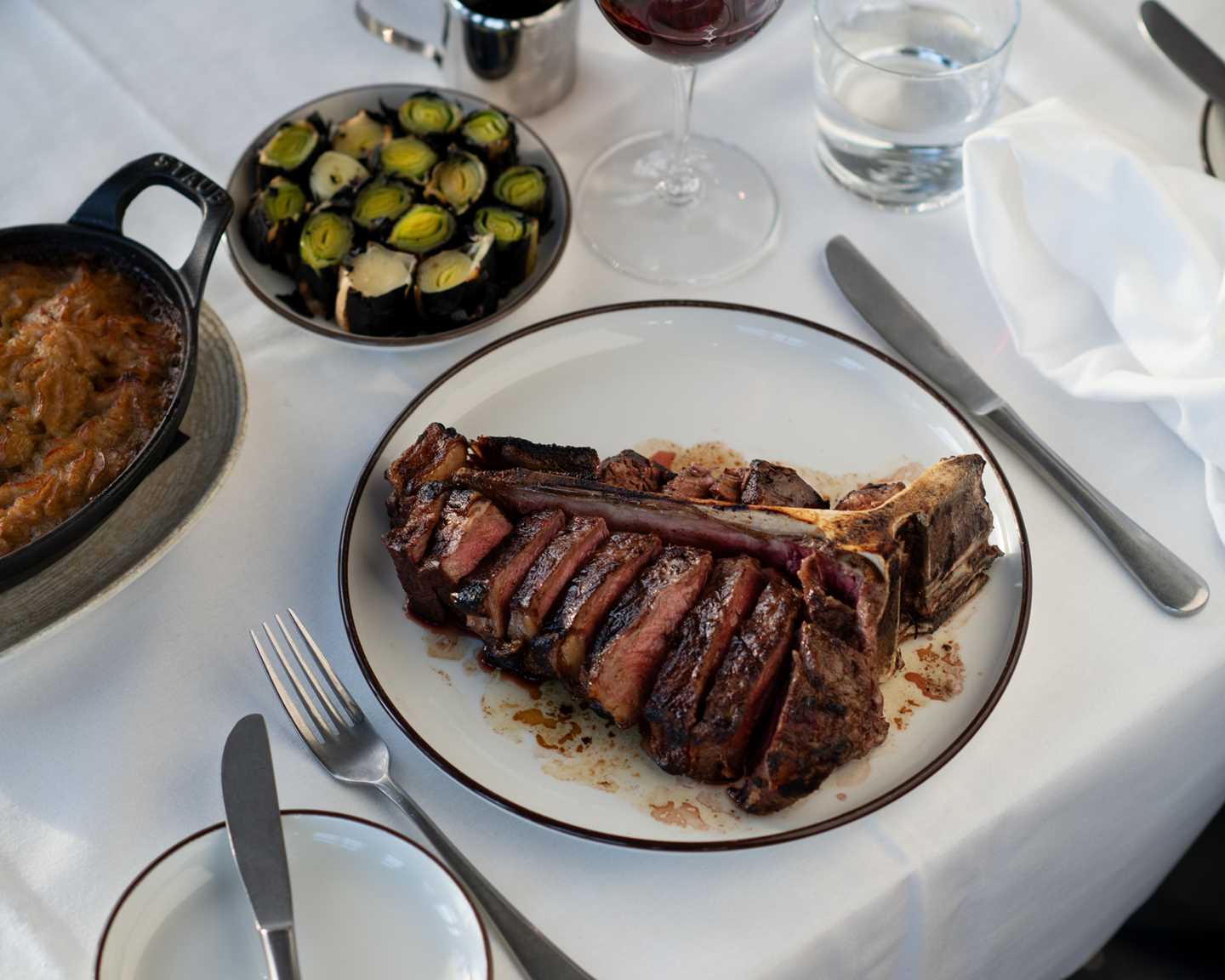 A table set with a white tablecloth, cutlery, and stemmed glasses. In focus is a plate with a hearty T-bone steak, beside a glass of red wine, a plate of roasted leeks, and a baking dish of potatoes. Photograph.