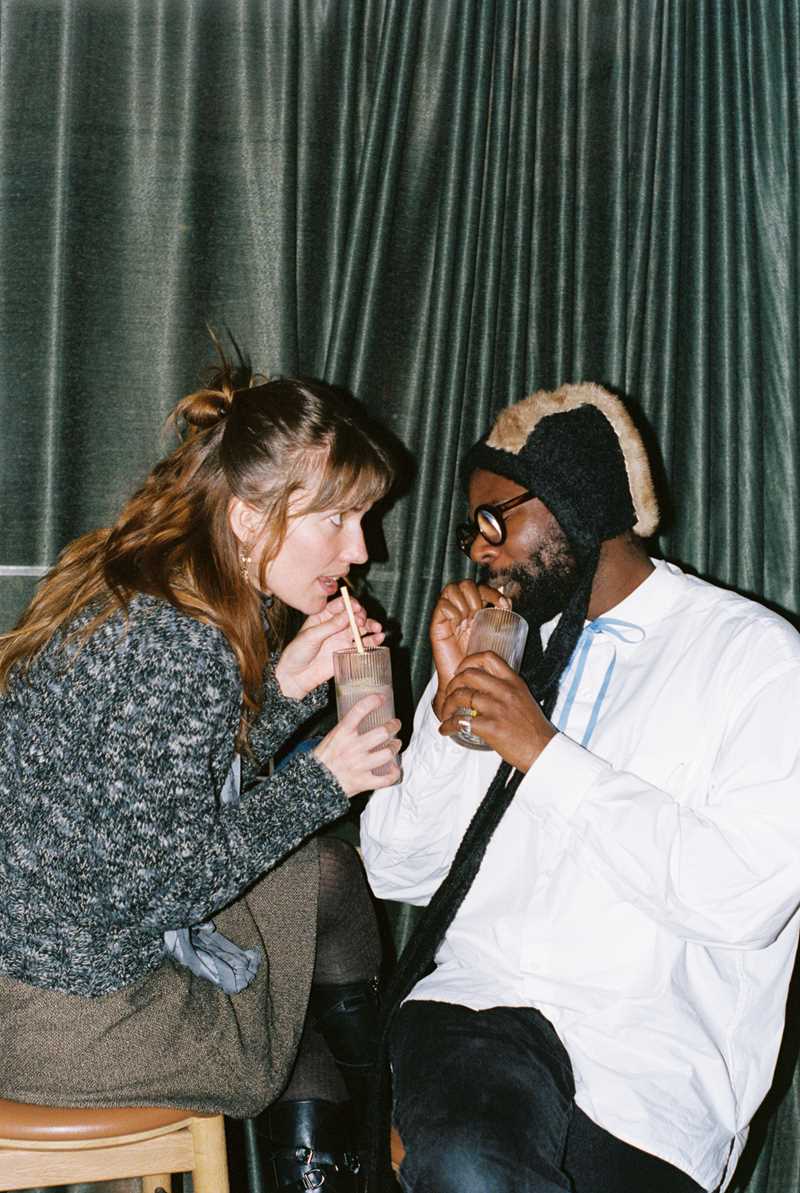 A woman and a man sit close, facing each other, drinking from their drinks with straws. The woman stares intently at the man, who has his eyes closed. Photograph.
