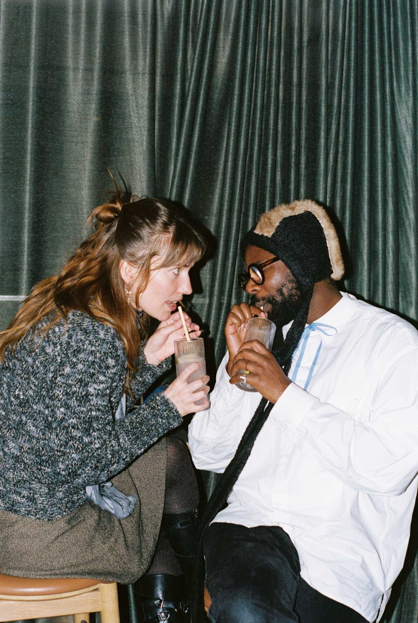 A woman and a man sit close, facing each other, drinking from their drinks with straws. The woman stares intently at the man, who has his eyes closed. Photograph.