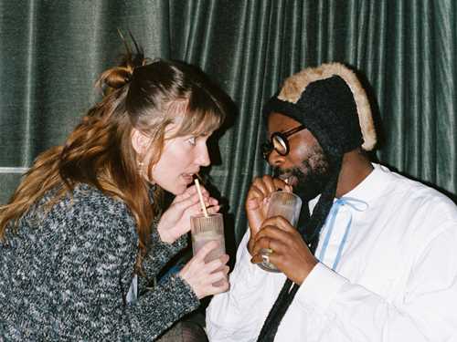 A woman and a man sit close, facing each other, drinking from their drinks with straws. The woman stares intently at the man, who has his eyes closed. Photograph.