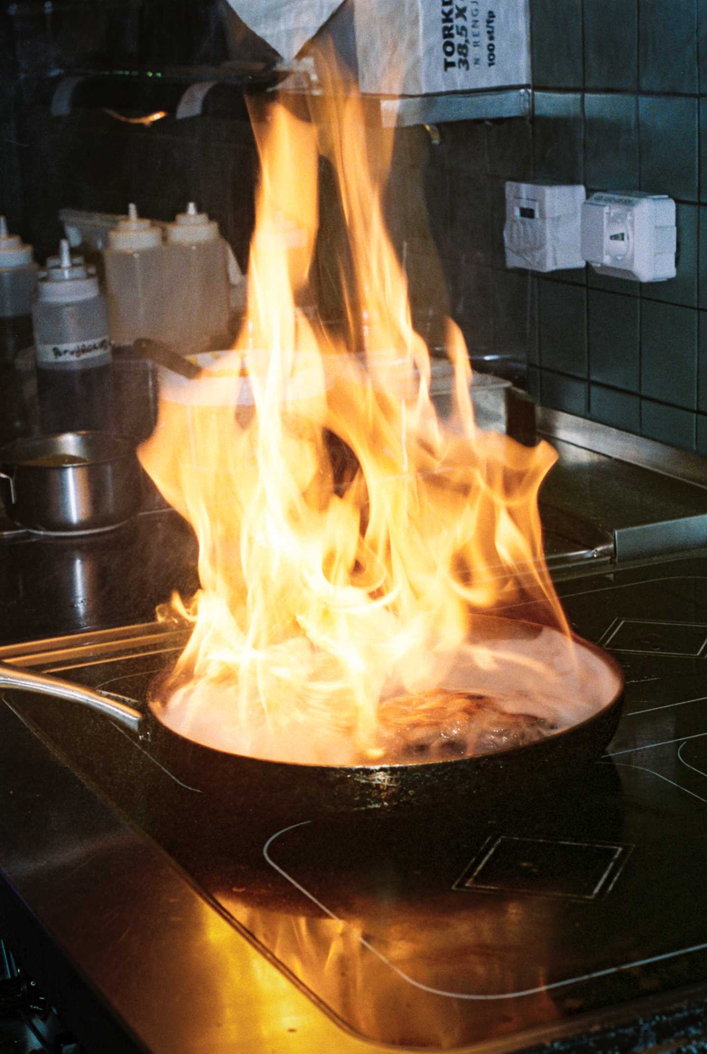 Large flames rise from a frying pan where something is being flambéed in a restaurant kitchen. Photograph.