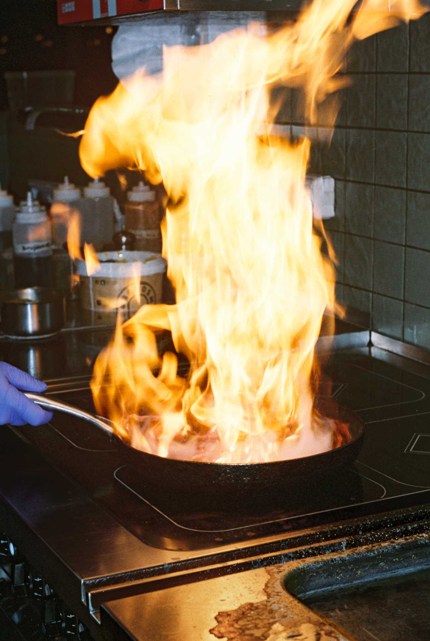 Large flames rise from a frying pan where something is being flambéed in a restaurant kitchen. A hand wearing a blue plastic glove holds the pan’s handle, and in the background, bottles of dressings, oils, and sauces are visible. Photograph.
