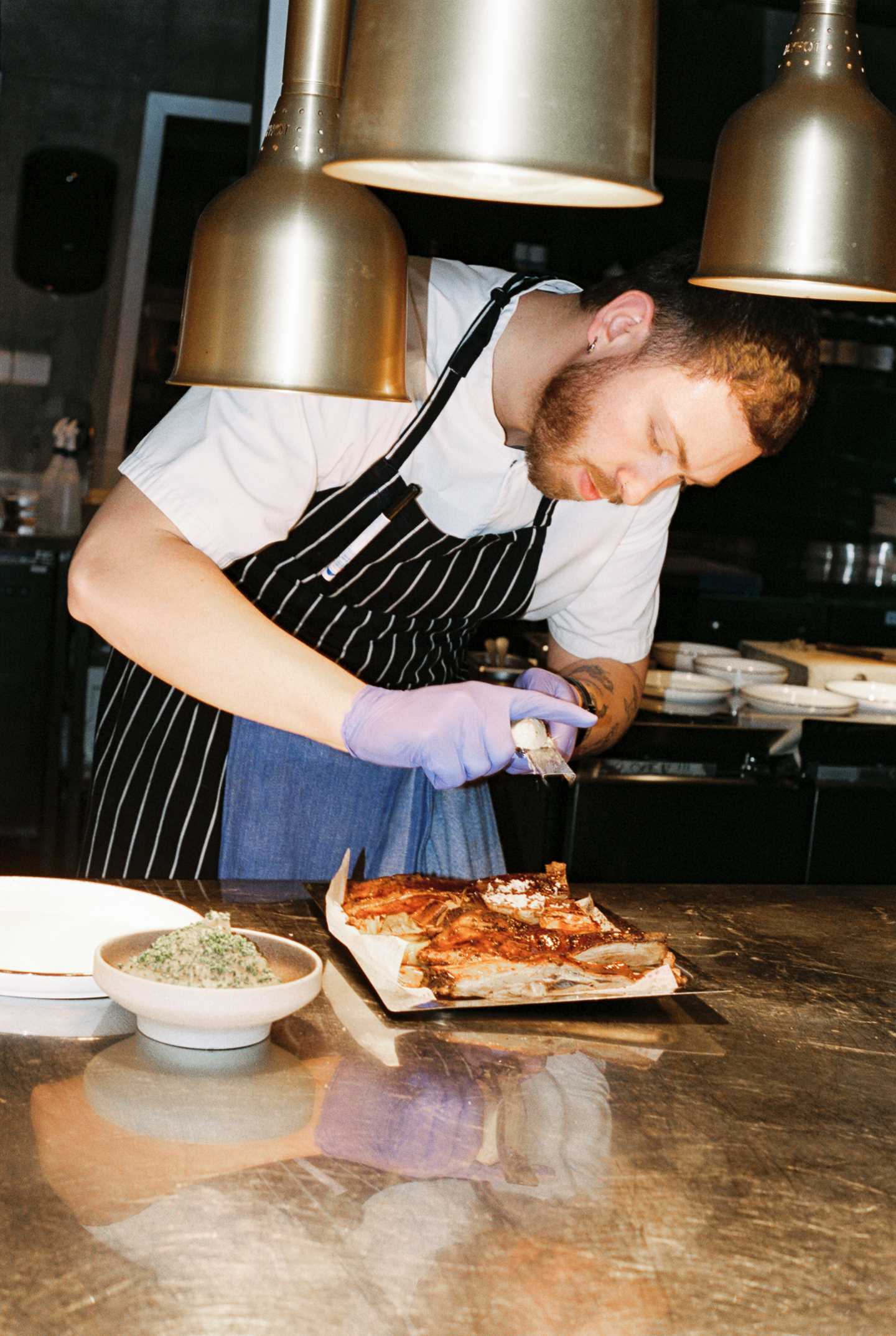 A chef leans over a dish, grating cheese on top. Heat lamps hang from the ceiling above and in front of him. Photograph.