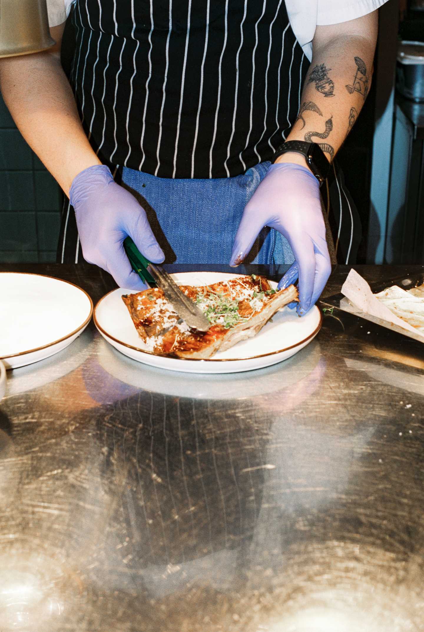 The arms of a chef wearing blue plastic gloves are visible as they place a slice of pizza on a plate. Photograph.