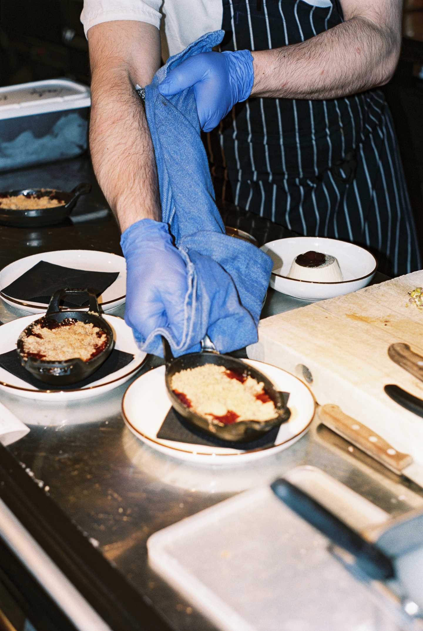 A chef wearing blue plastic gloves and a striped apron lifts a baking dish from the oven onto a plate, using a kitchen towel to avoid burning himself. Photograph.