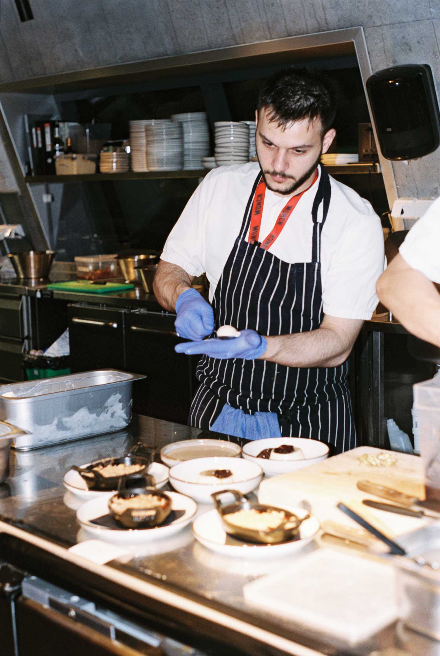 A chef wearing a striped apron and blue plastic gloves shapes elongated scoops of ice cream in their hand. Photograph.