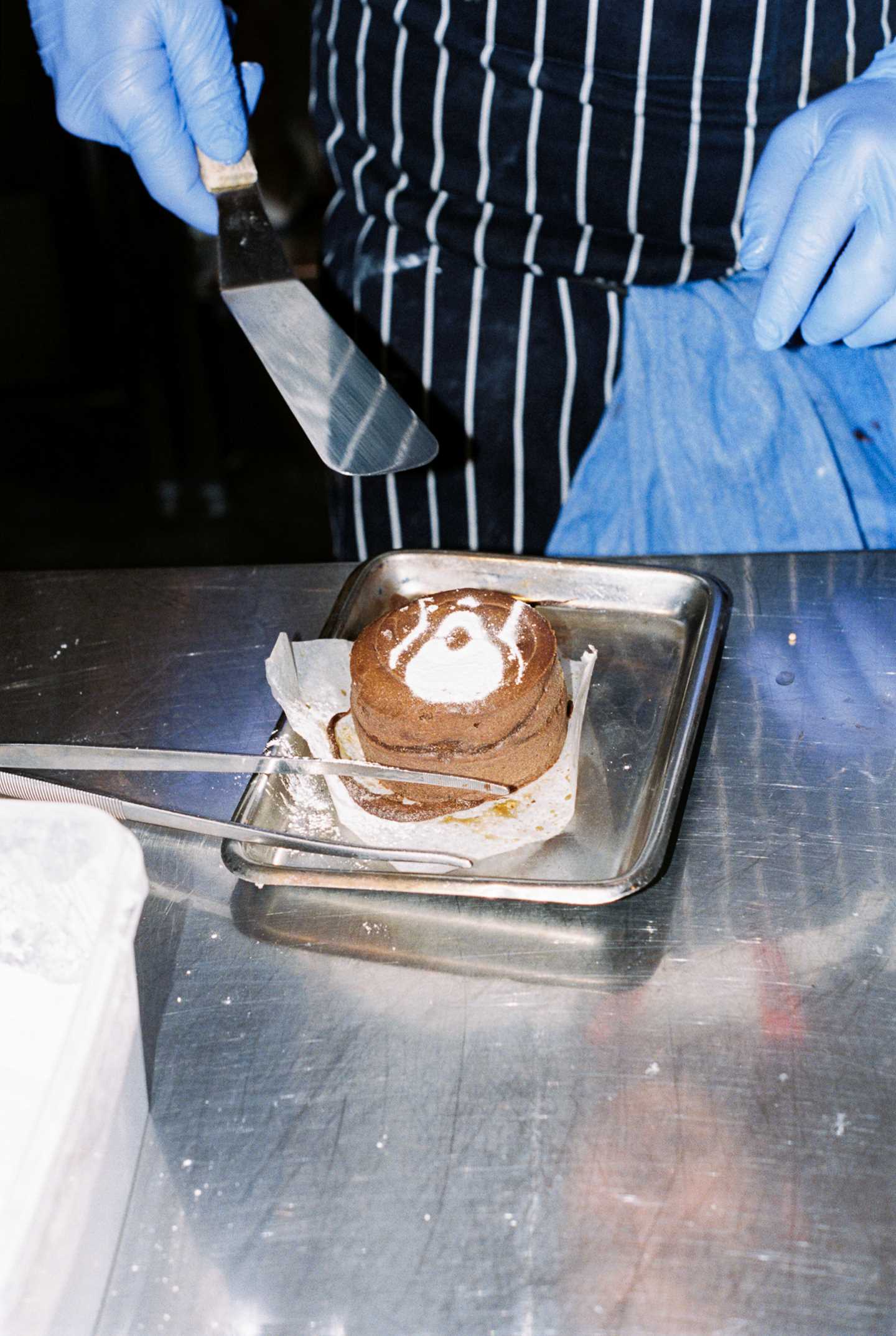 A chocolate fondant decorated with powdered sugar in the shape of The Scream motif. In the background, a chef’s hands holding a spatula are visible. Photograph.