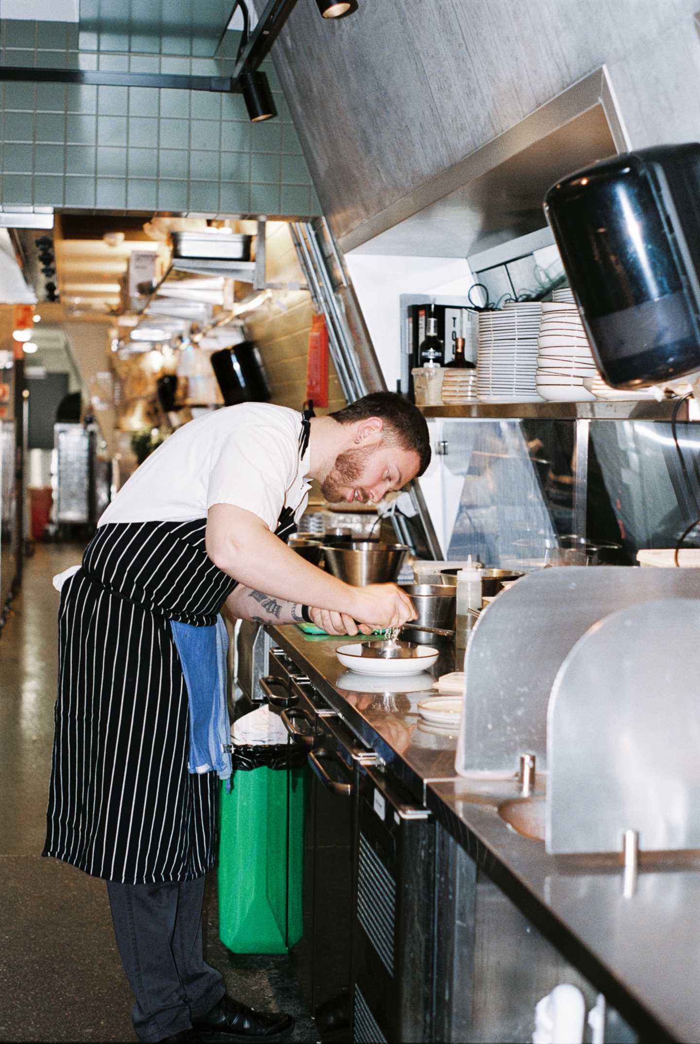 A chef leans over a kitchen counter in a restaurant kitchen, grating something white over a dish. Photograph.