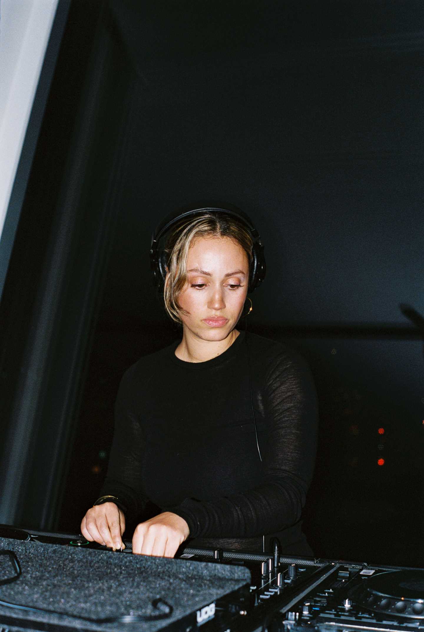 A DJ dressed in black with blonde hair and wearing headphones stands behind a mixing console in front of a large window. Outside, the sky is pitch black. Photograph.