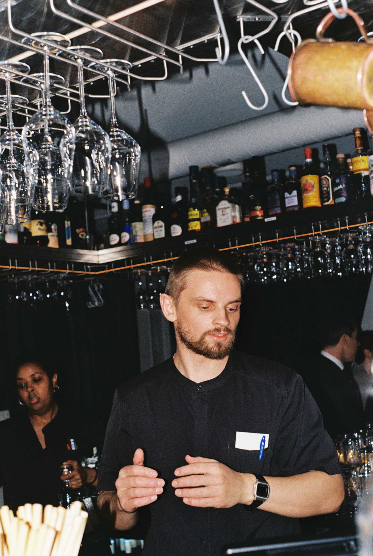 A bartender stands in the centre of a square bar counter. On a shelf near the ceiling are various bottles of spirits, and stemmed glasses hang upside down from racks beneath the shelf. Photograph.
